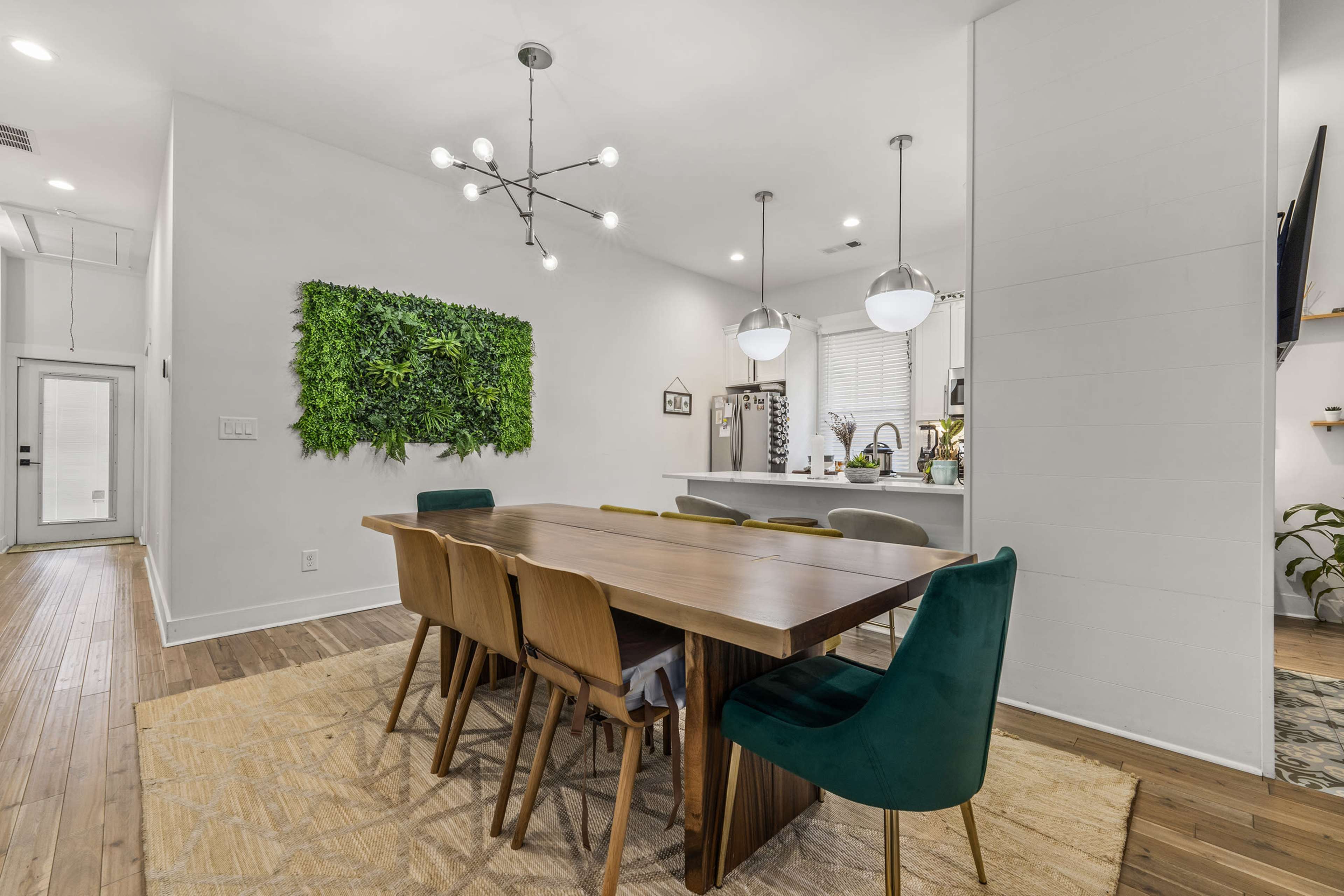A dining area features a large wooden table surrounded by a mix of green and neutral chairs, with a wall of greenery and modern light fixtures above.