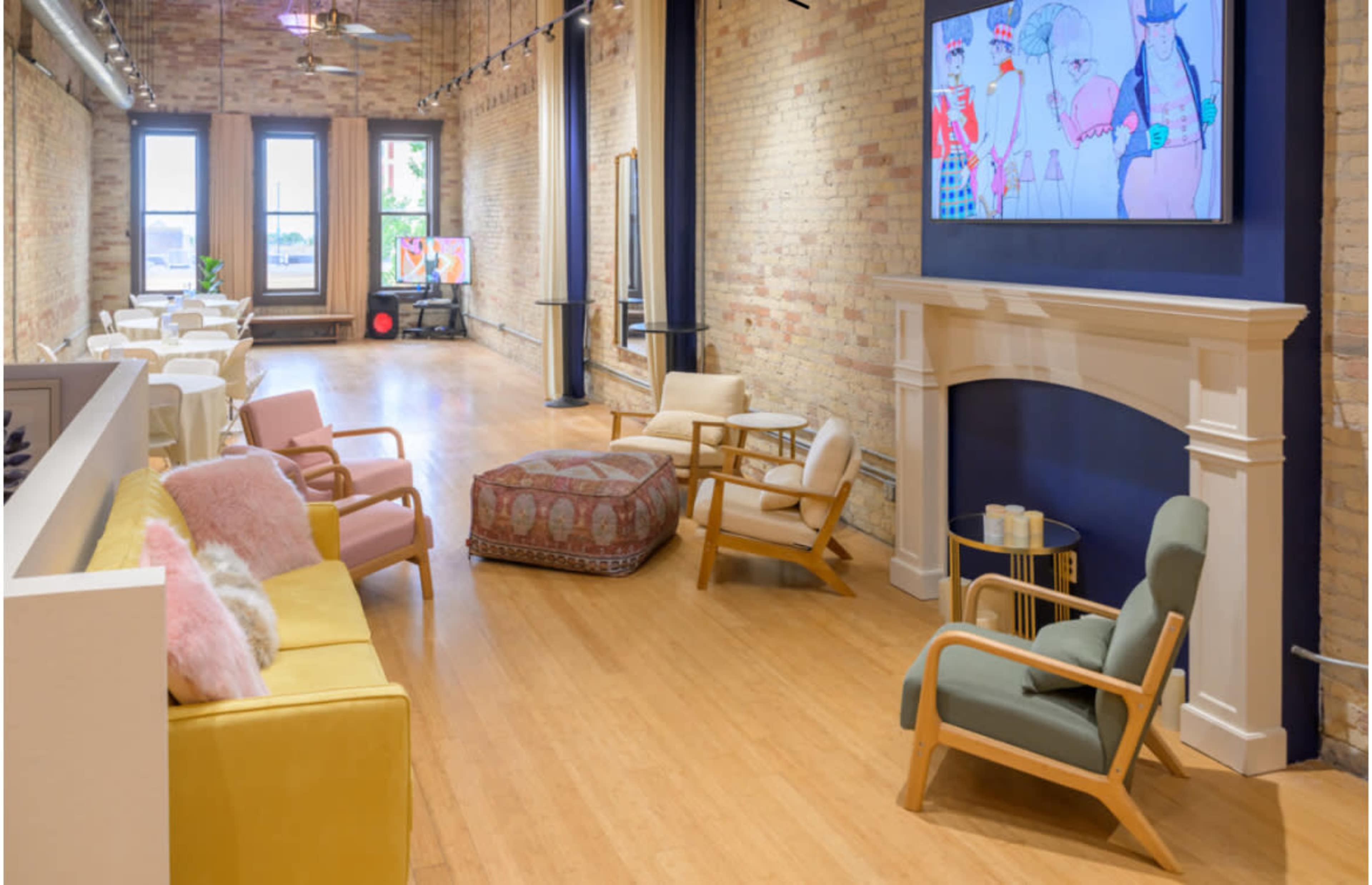 The image shows a spacious interior with a blend of seating options, including colorful couches and armchairs, arranged in a well-lit room featuring exposed brick walls and artistic decor.