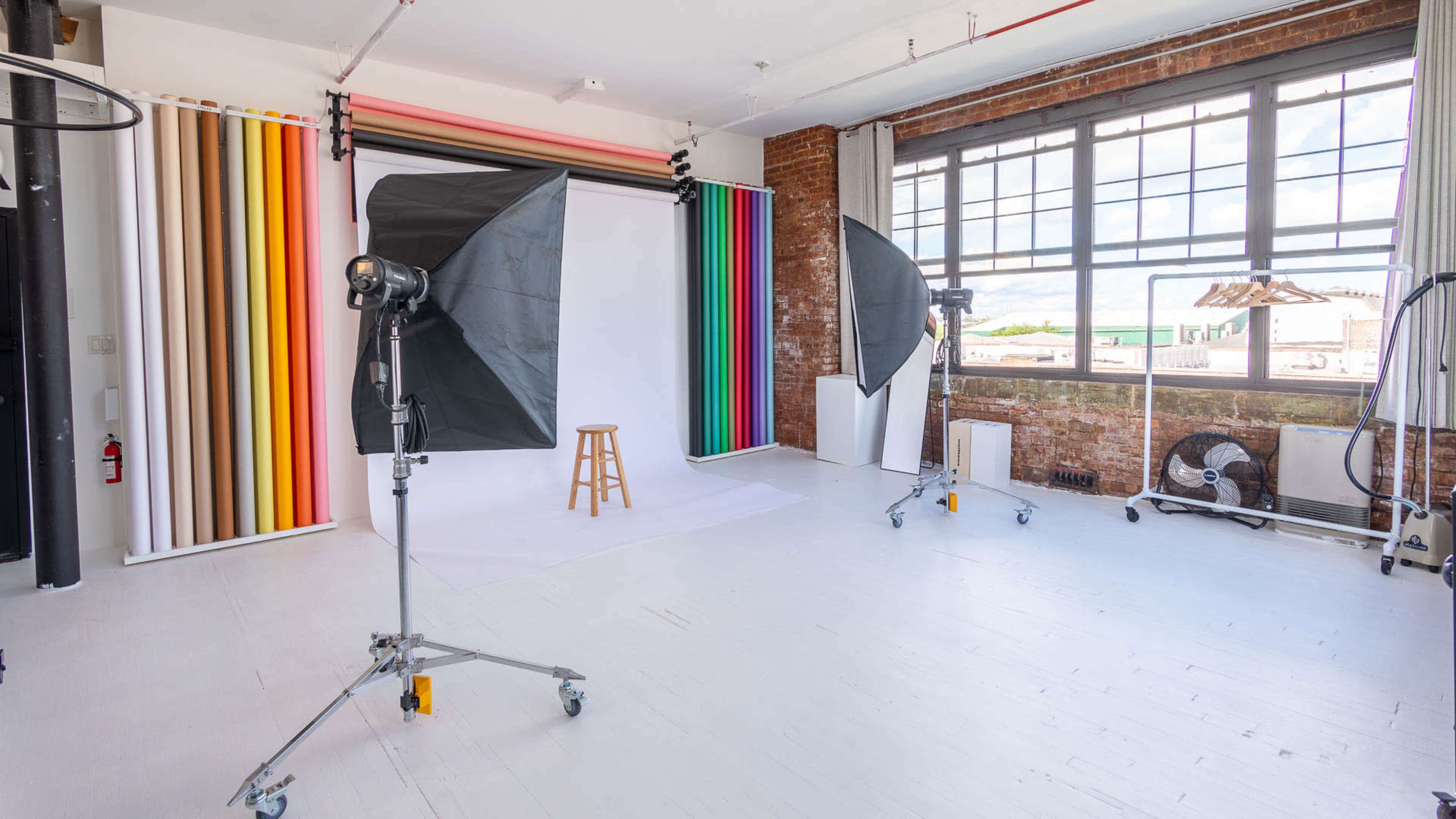 A photography studio with a stool, backdrop, lighting equipment, and large windows.