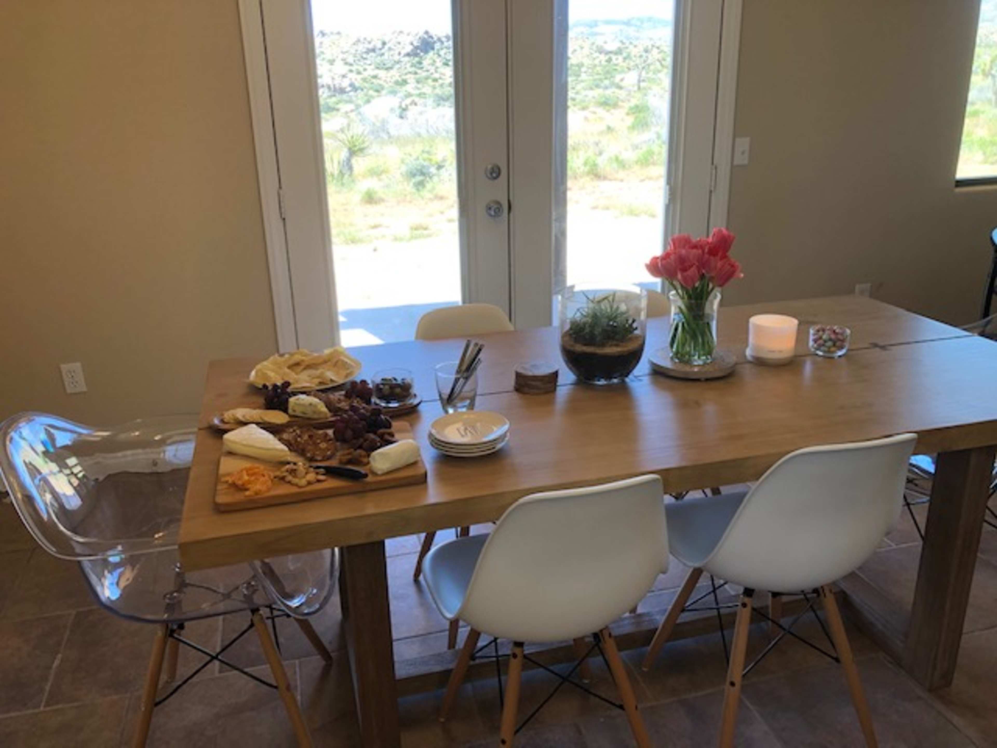 A wooden dining table is set with a variety of cheeses, fruits, and flowers, while large glass doors provide a view of the outdoors.