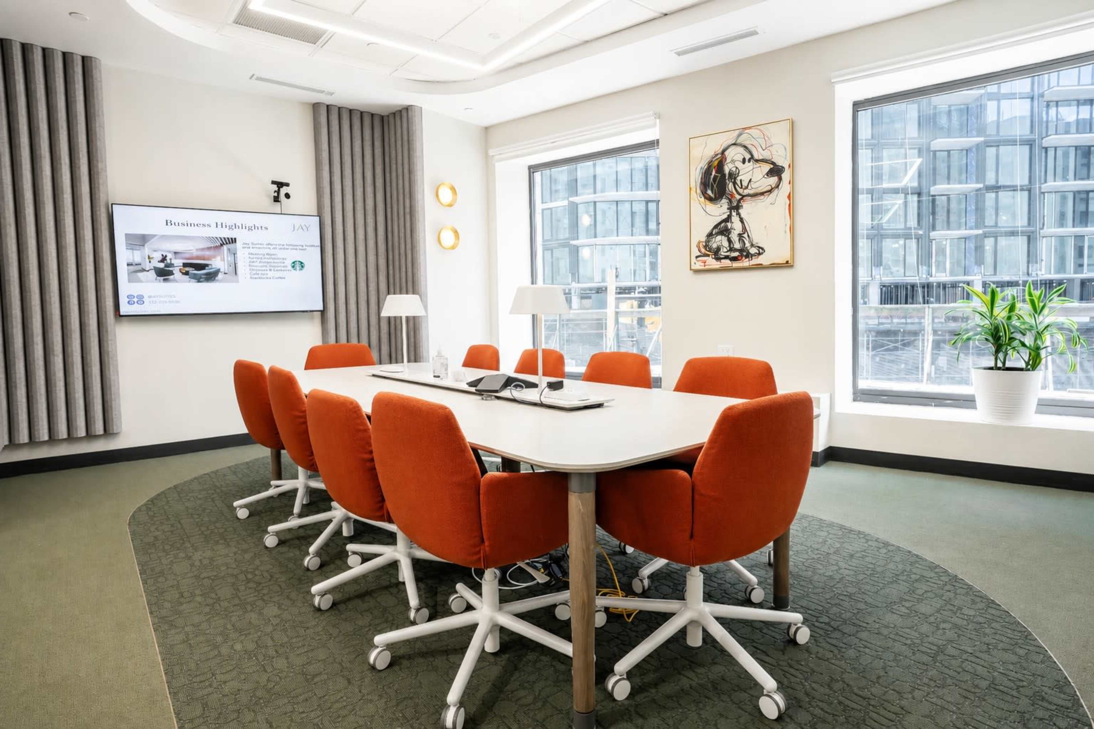 A modern conference room features a large table surrounded by orange swivel chairs, with a screen displaying business highlights and large windows letting in natural light.