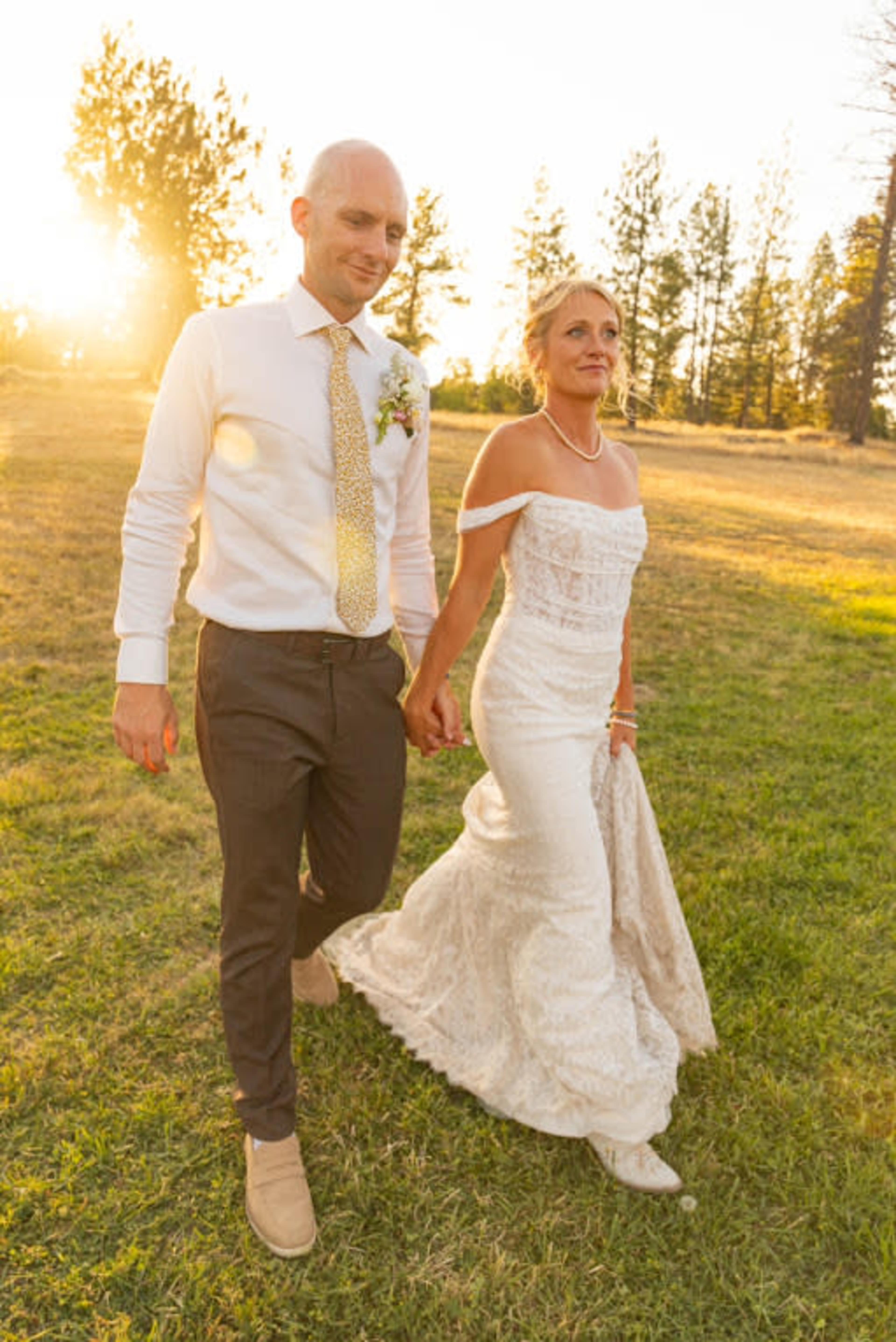 A bride and groom walk hand in hand through a grassy area with trees and a sunset in the background.