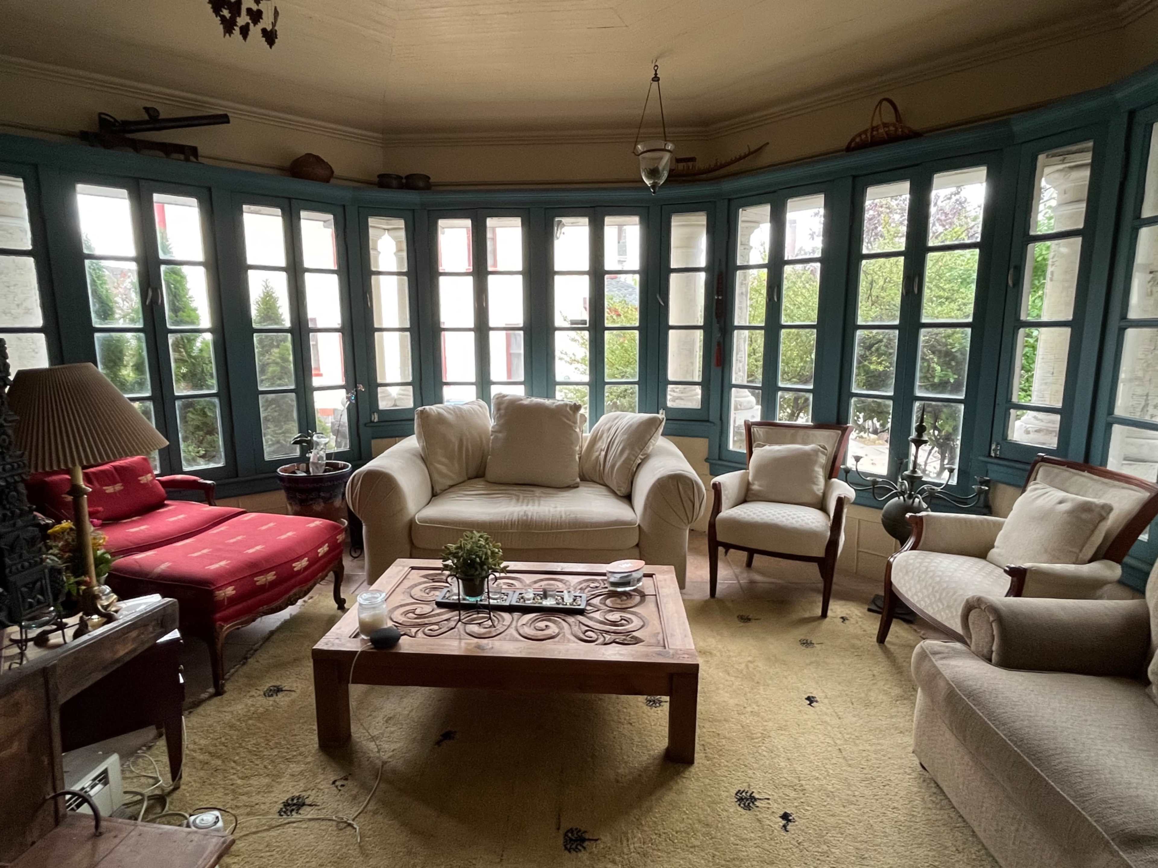 A cozy sunroom with large windows, a wooden coffee table, and several armchairs arranged around a central seating area.