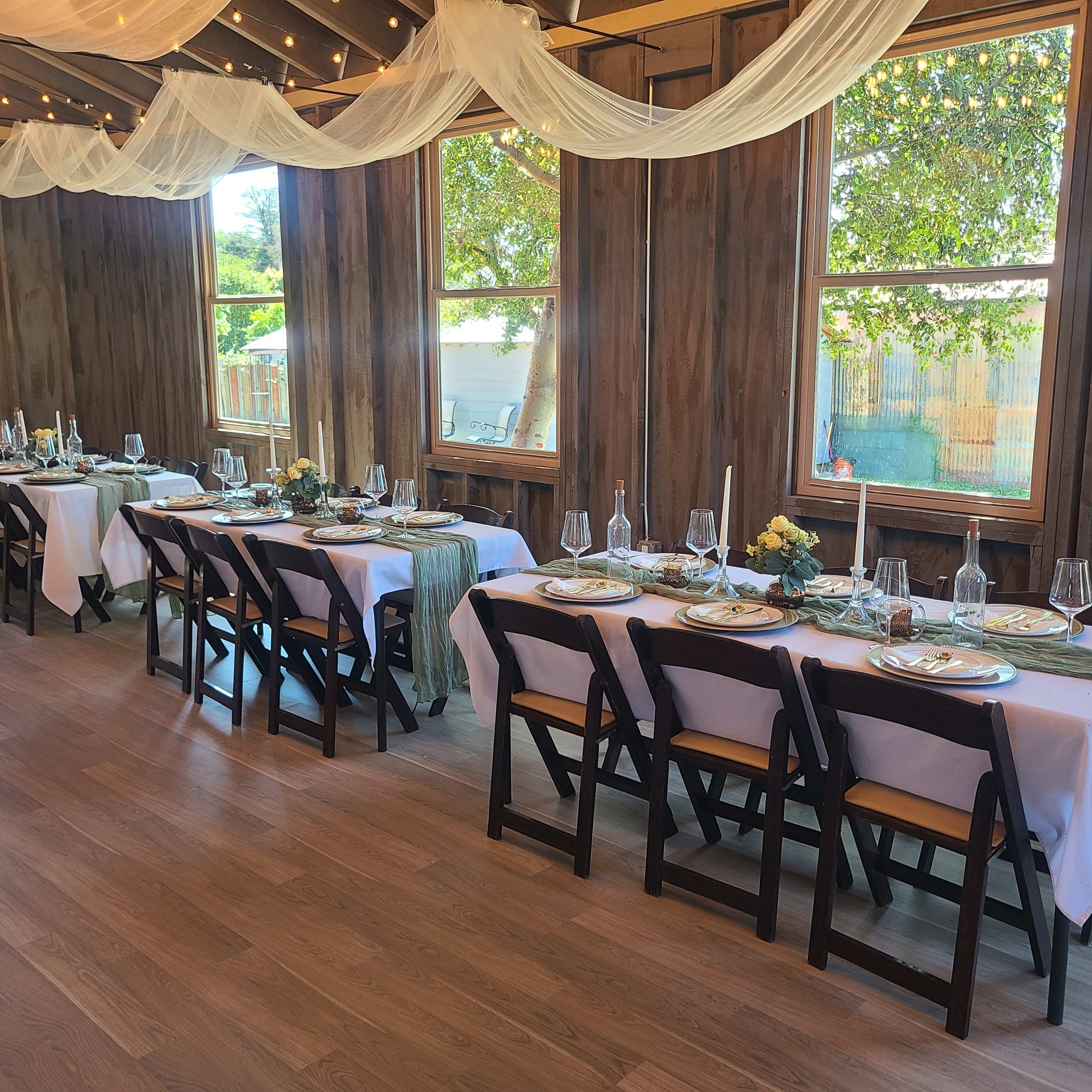 A long, elegantly set dining table is arranged along the wooden walls of a bright room with large windows.