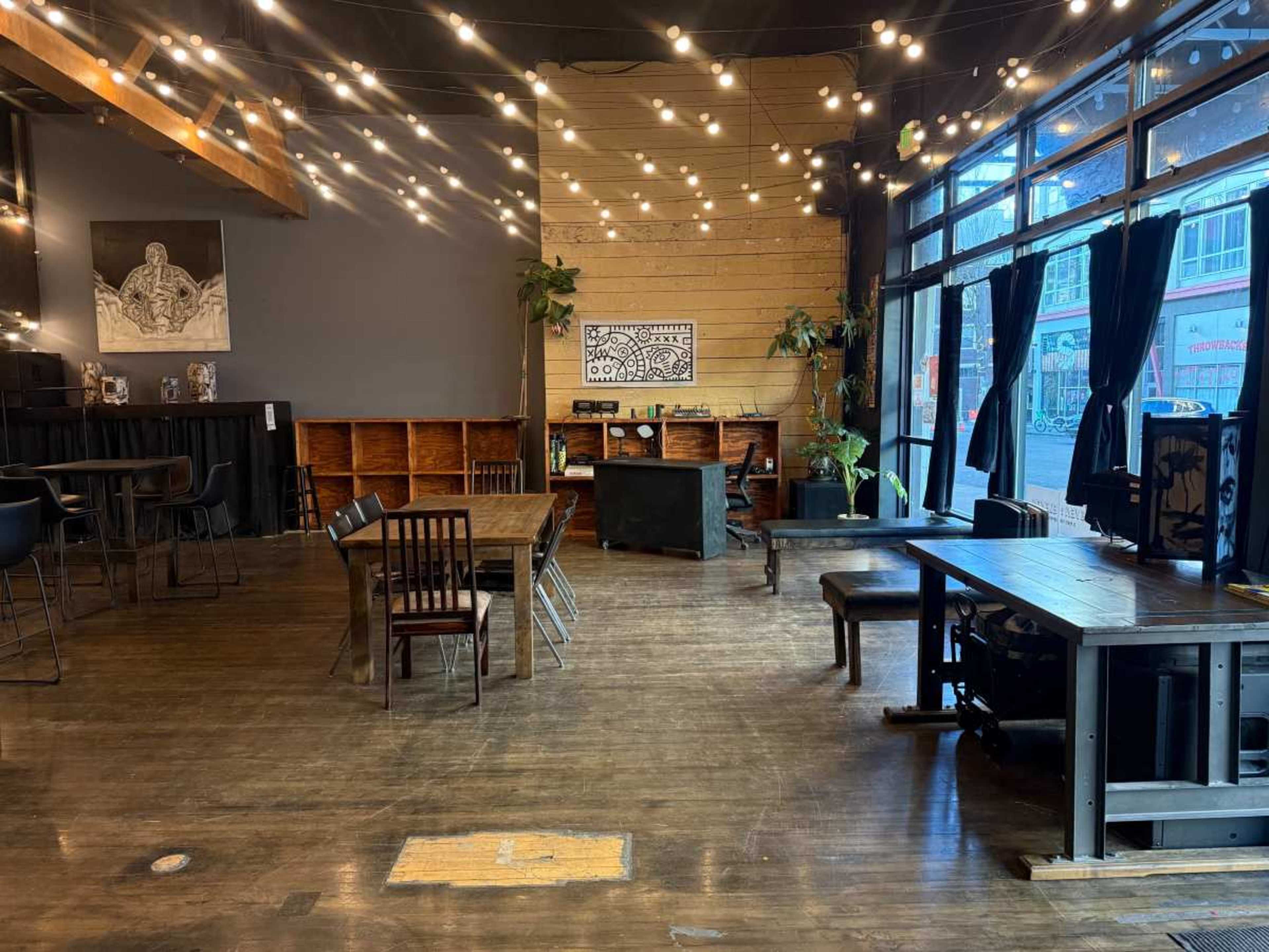The image shows a spacious, well-lit café or event space with wooden floors, tables, and string lights hanging overhead.