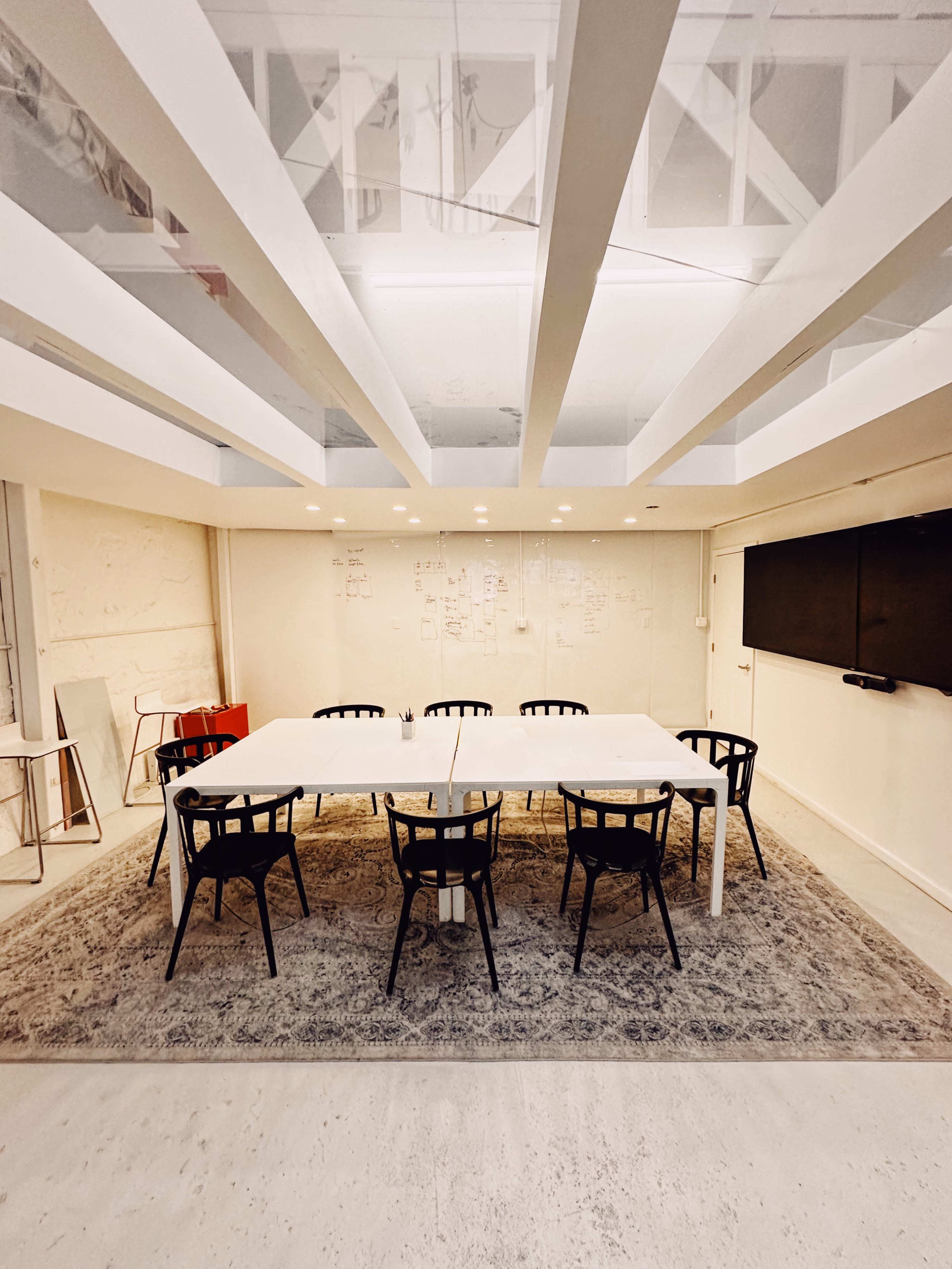 A modern conference room features a long table surrounded by black chairs on a light-colored rug, with a whiteboard and wall-mounted screen in the background.