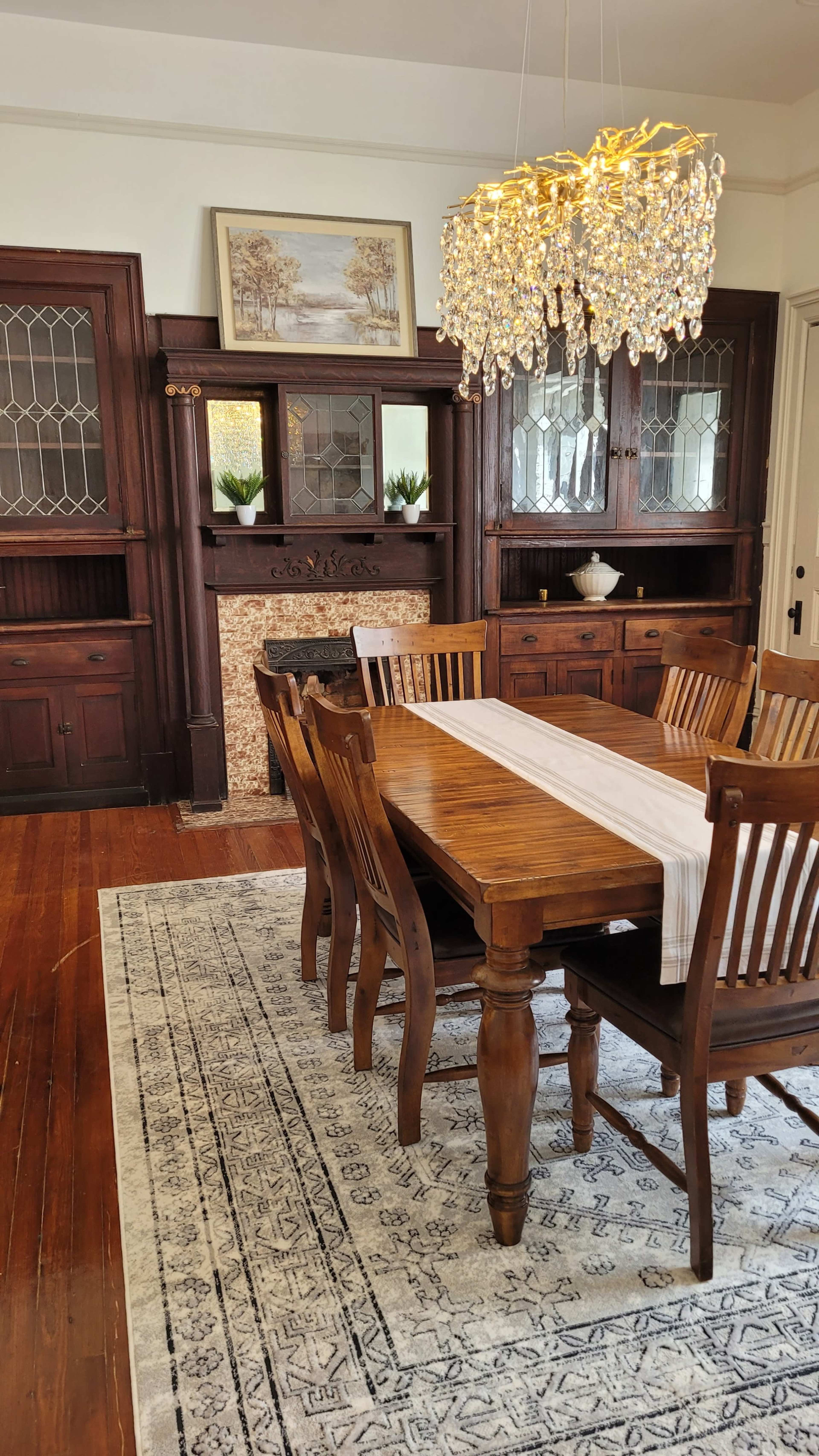 A dining room with a wooden table surrounded by six chairs, a patterned rug on the floor, and a chandelier hanging above the table.