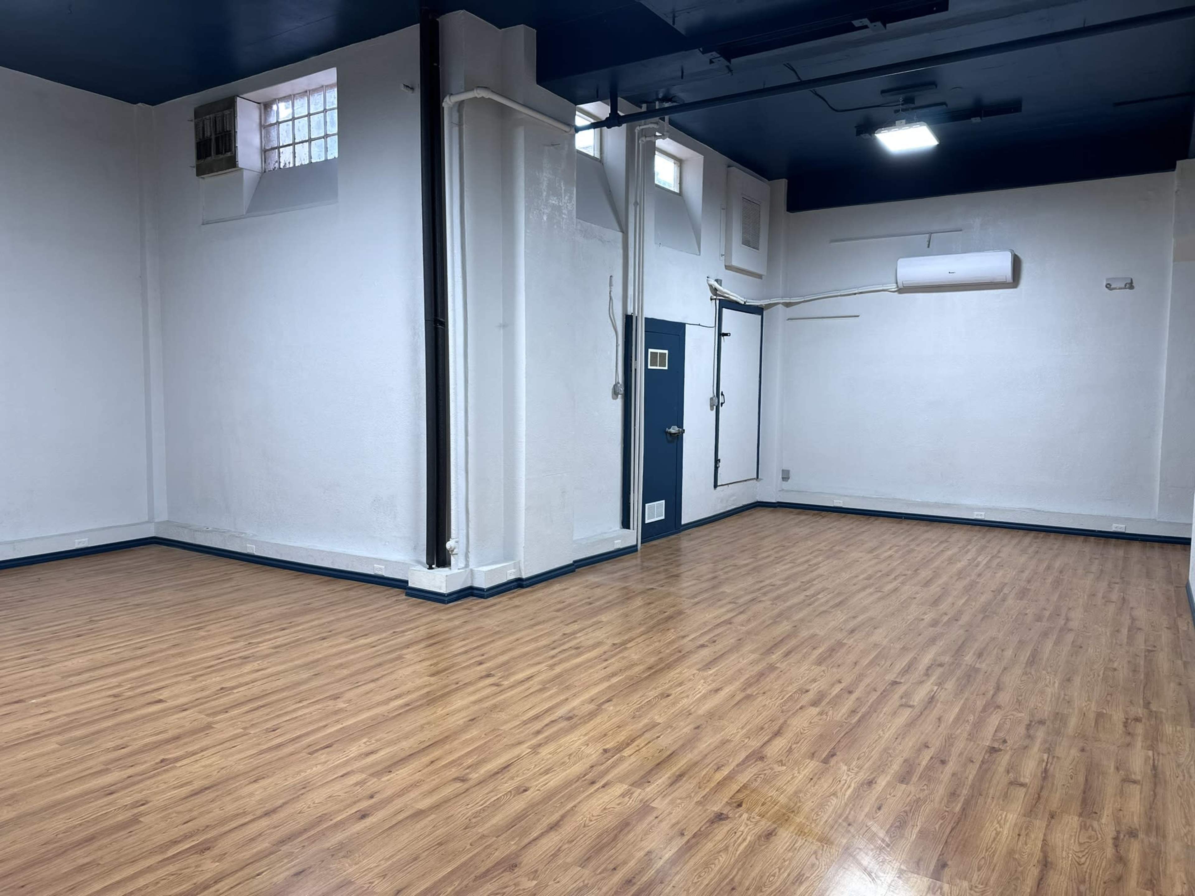 A spacious, empty room with wooden floors and plain white walls, featuring a single door and a window.