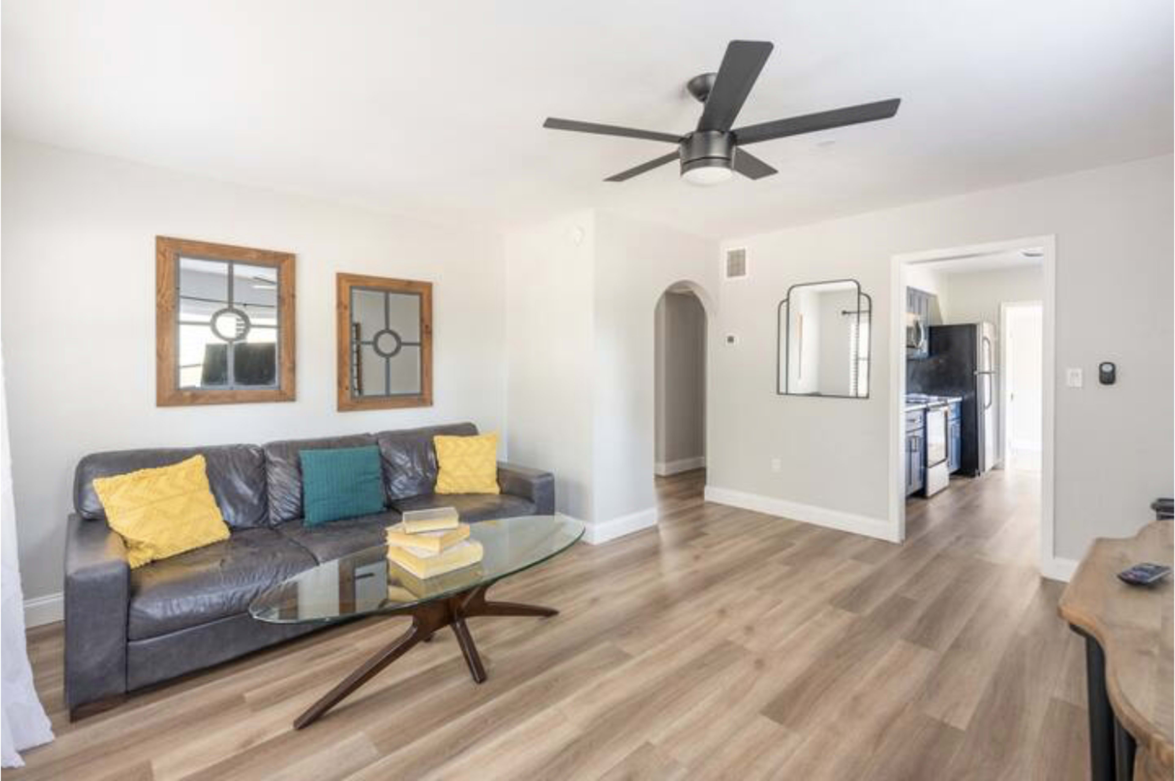 A living room features a dark leather sofa with yellow pillows, a glass coffee table, and two framed mirrors on the wall, with a doorway leading to the kitchen area.