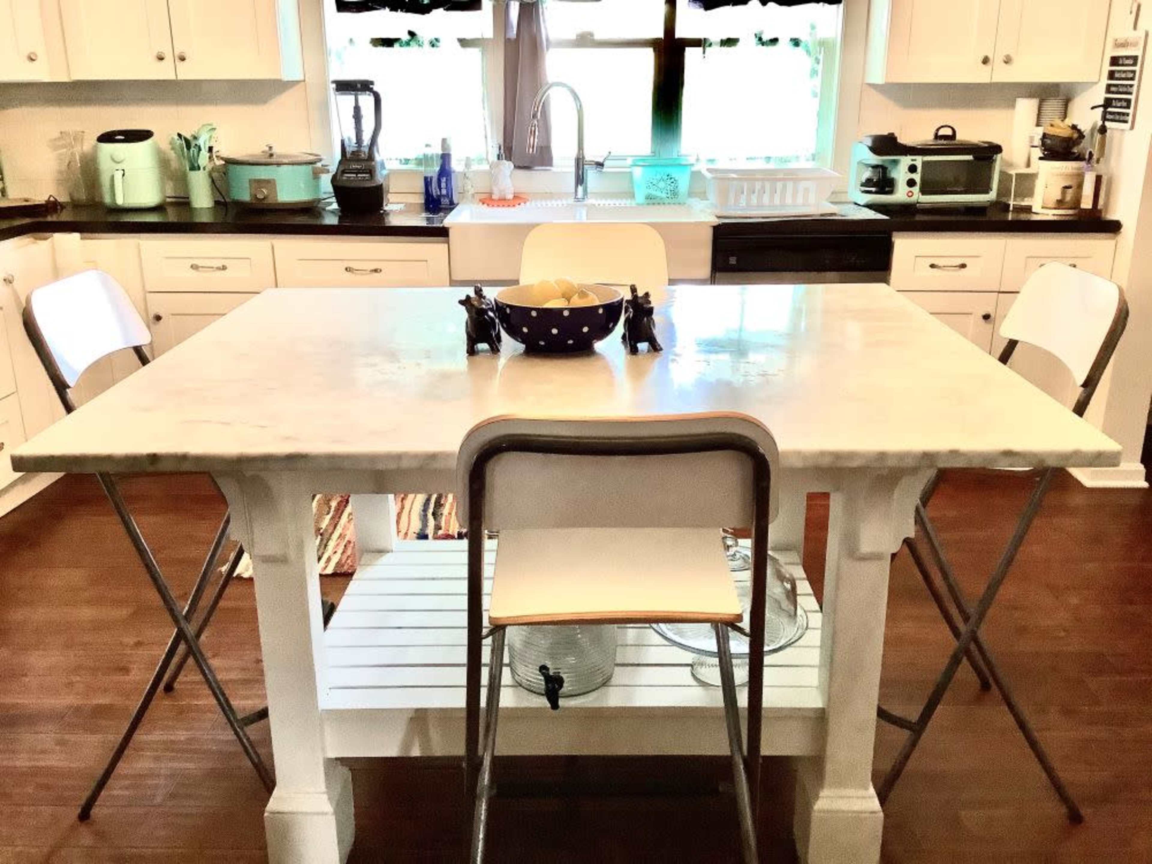 A kitchen featuring a marble-topped table with two folding chairs, surrounded by white cabinetry and various appliances.