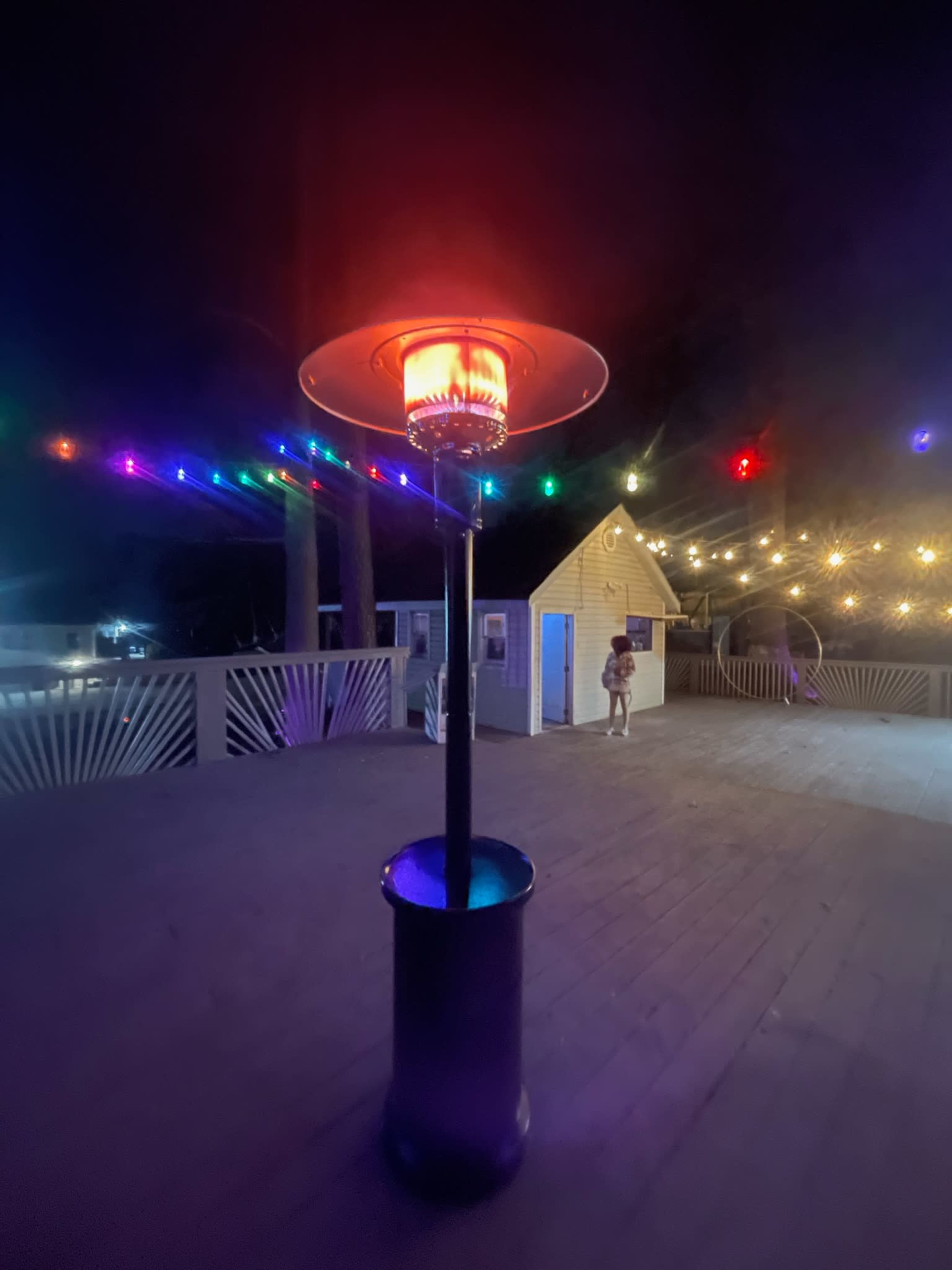 A glowing patio heater stands in the foreground, surrounded by colorful string lights illuminating a wooden deck at night.