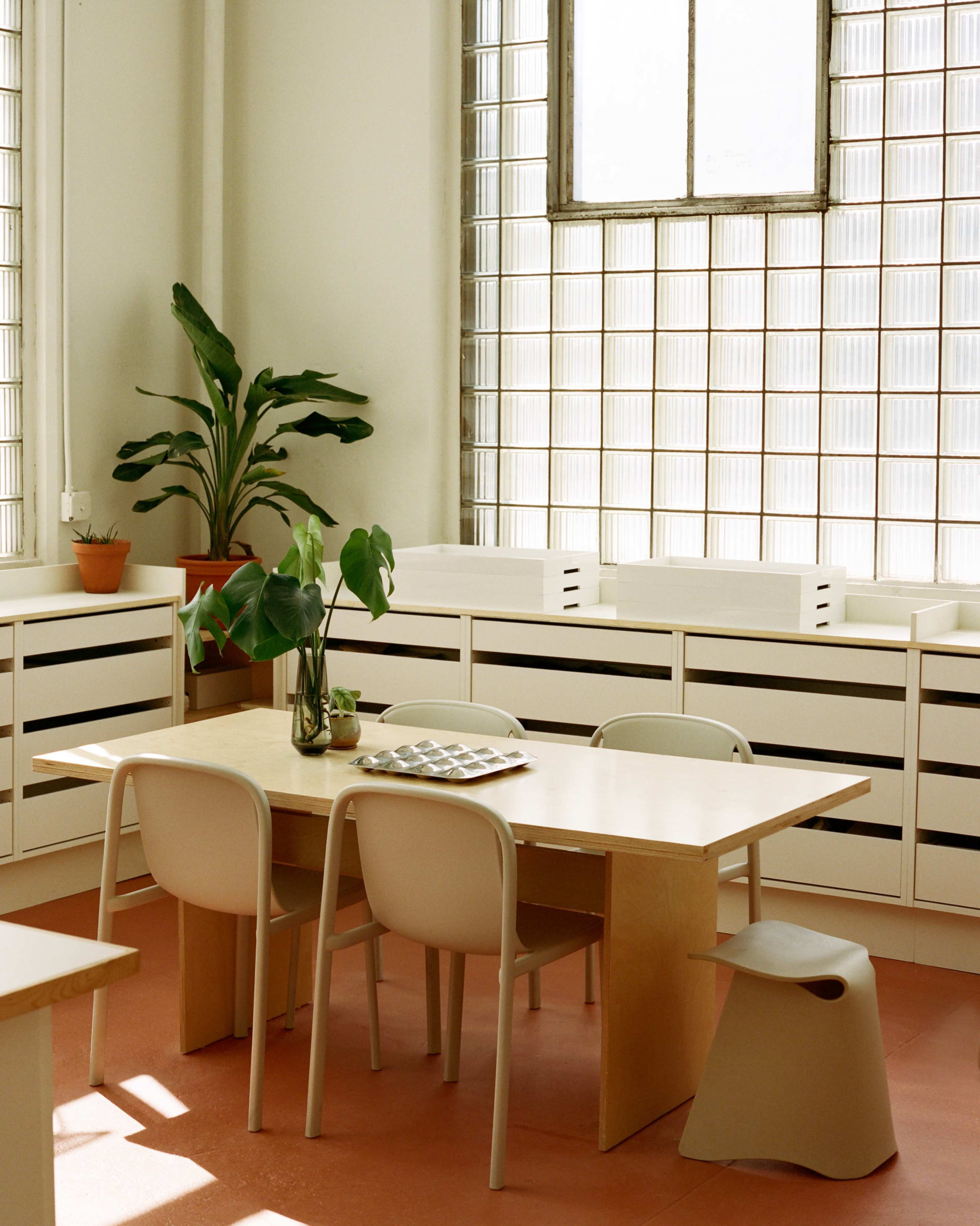 The image shows a bright, minimalist room with a wooden table and chairs, a potted plant, and large windows made of glass blocks.