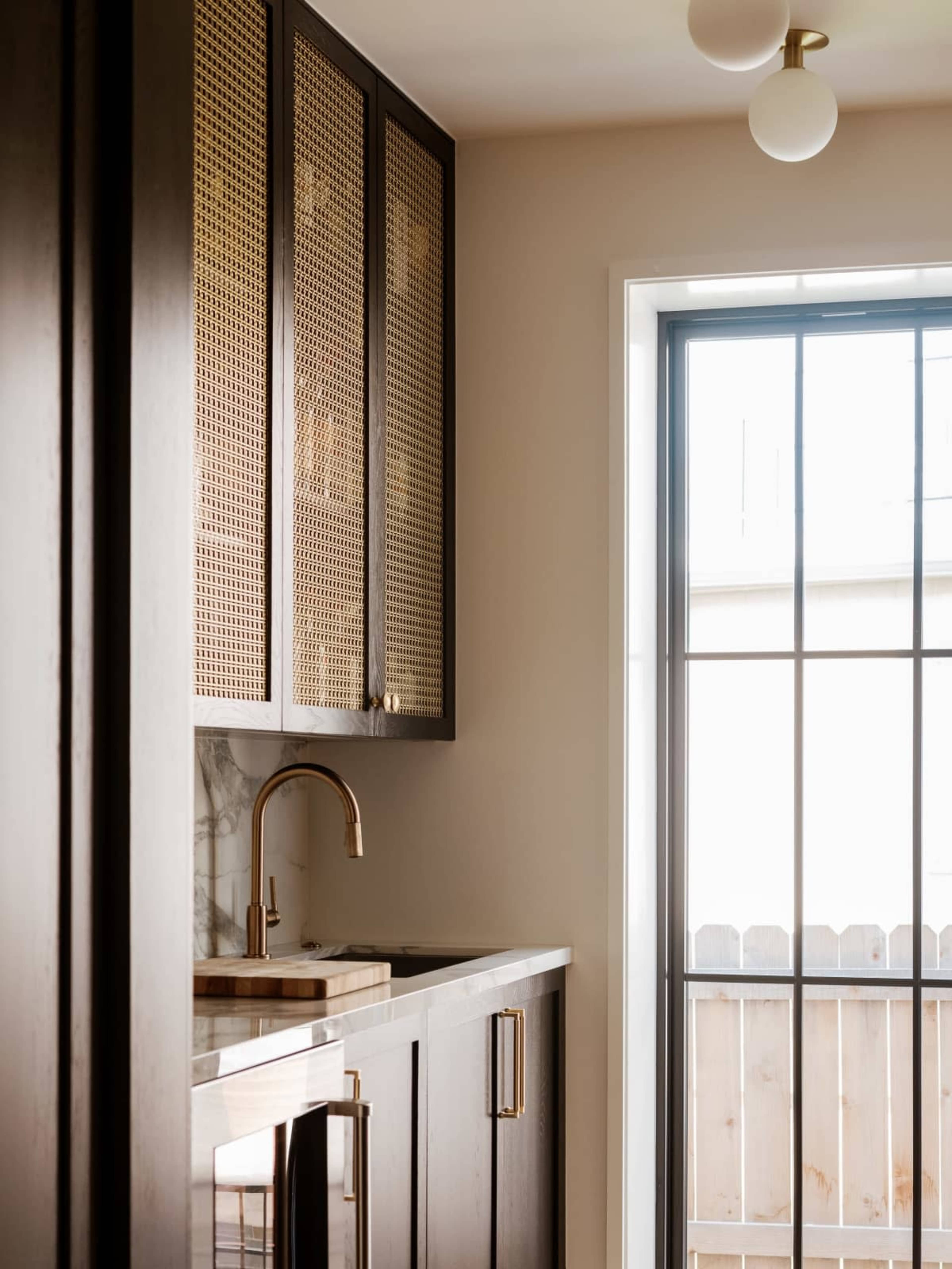 The image shows a modern kitchen with dark cabinetry, woven rattan cabinet doors, a marble countertop, and a window with a grid design.