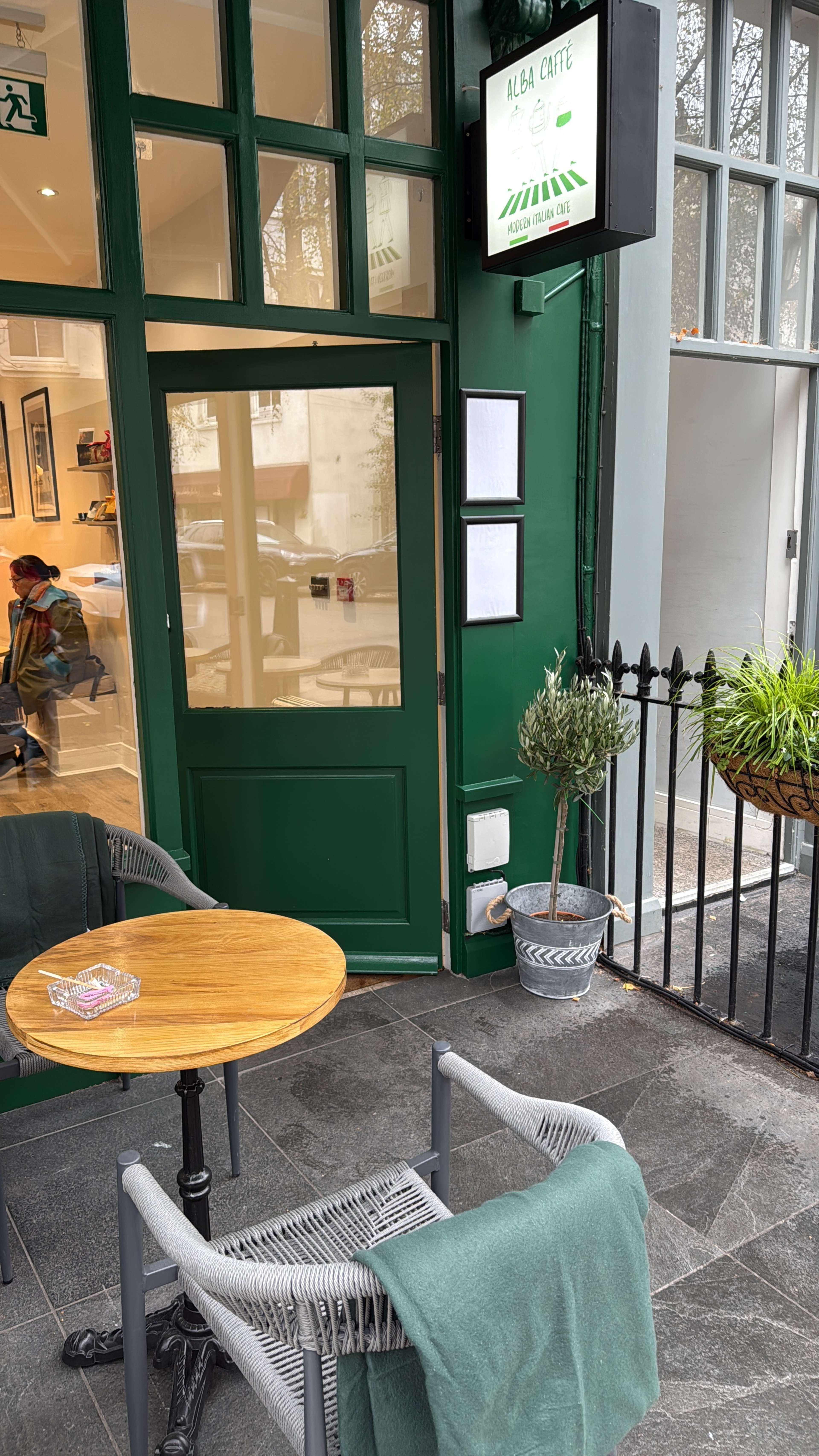 A small outdoor seating area features a wooden table and a chair with a green blanket, beside a green wall and a café sign.
