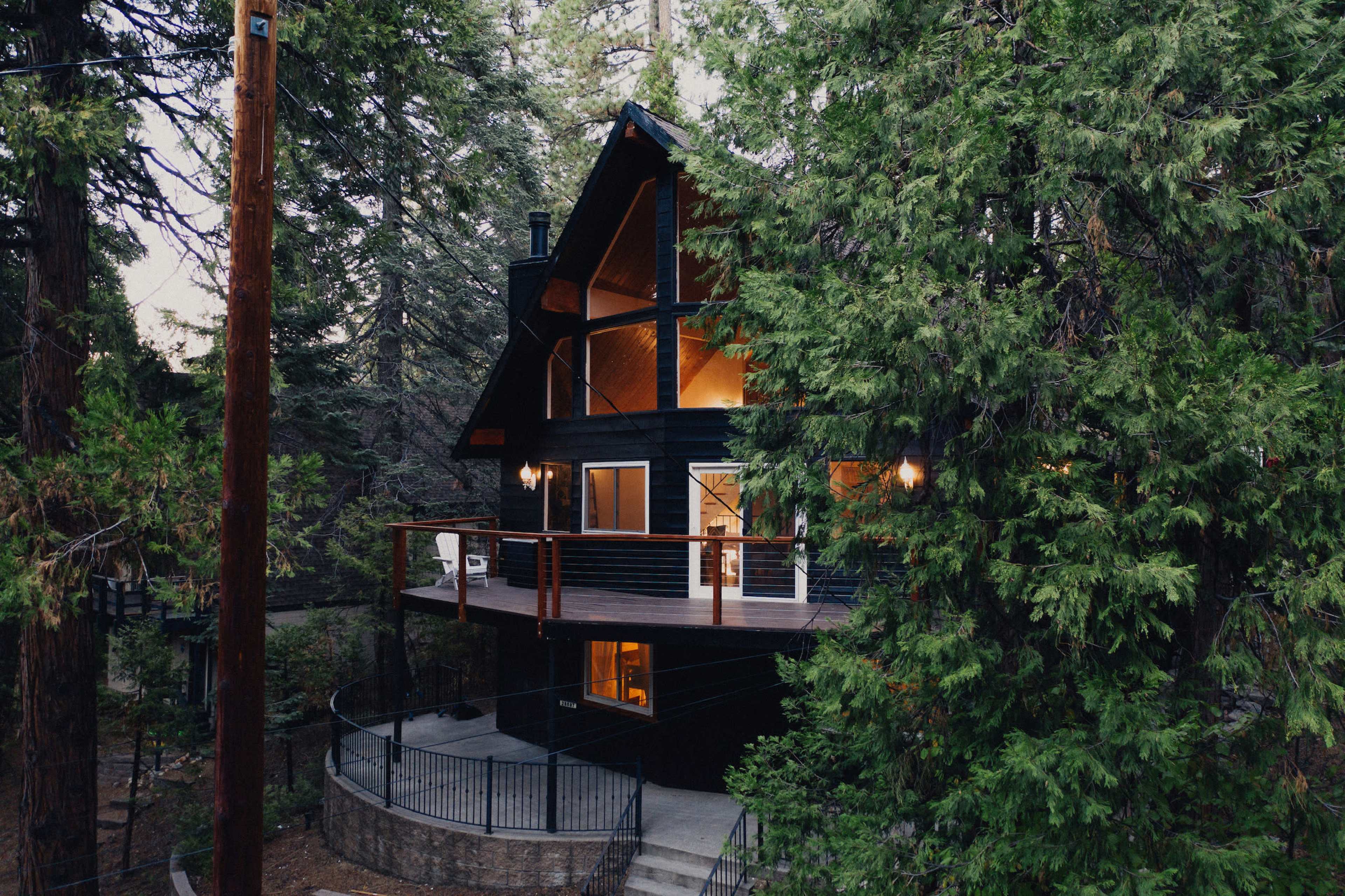 A black wooden cabin with a balcony is nestled among tall green trees.