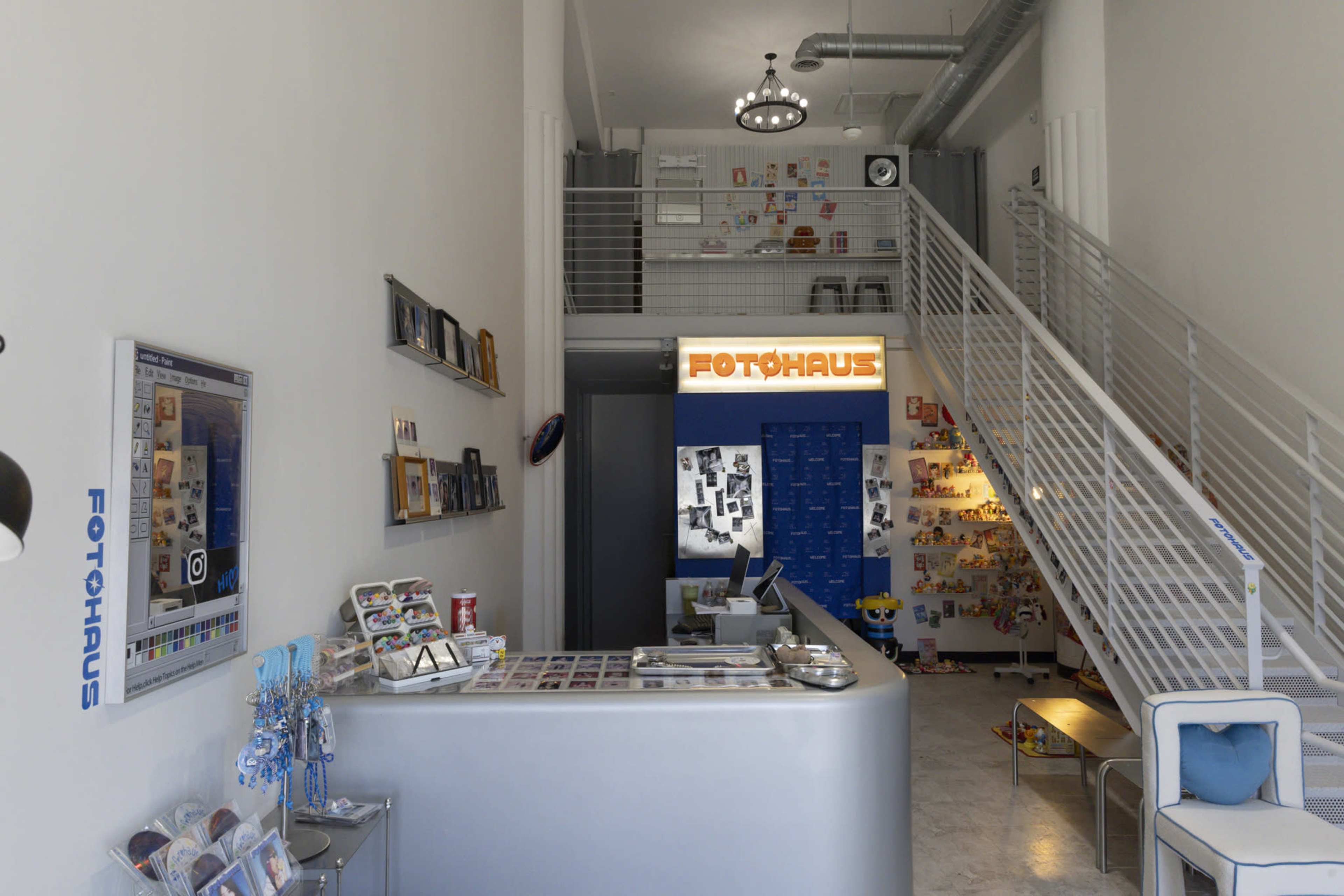 The image shows a modern photo studio reception area with a sleek counter, a wall displaying various framed pictures, and a staircase leading to an upper level.