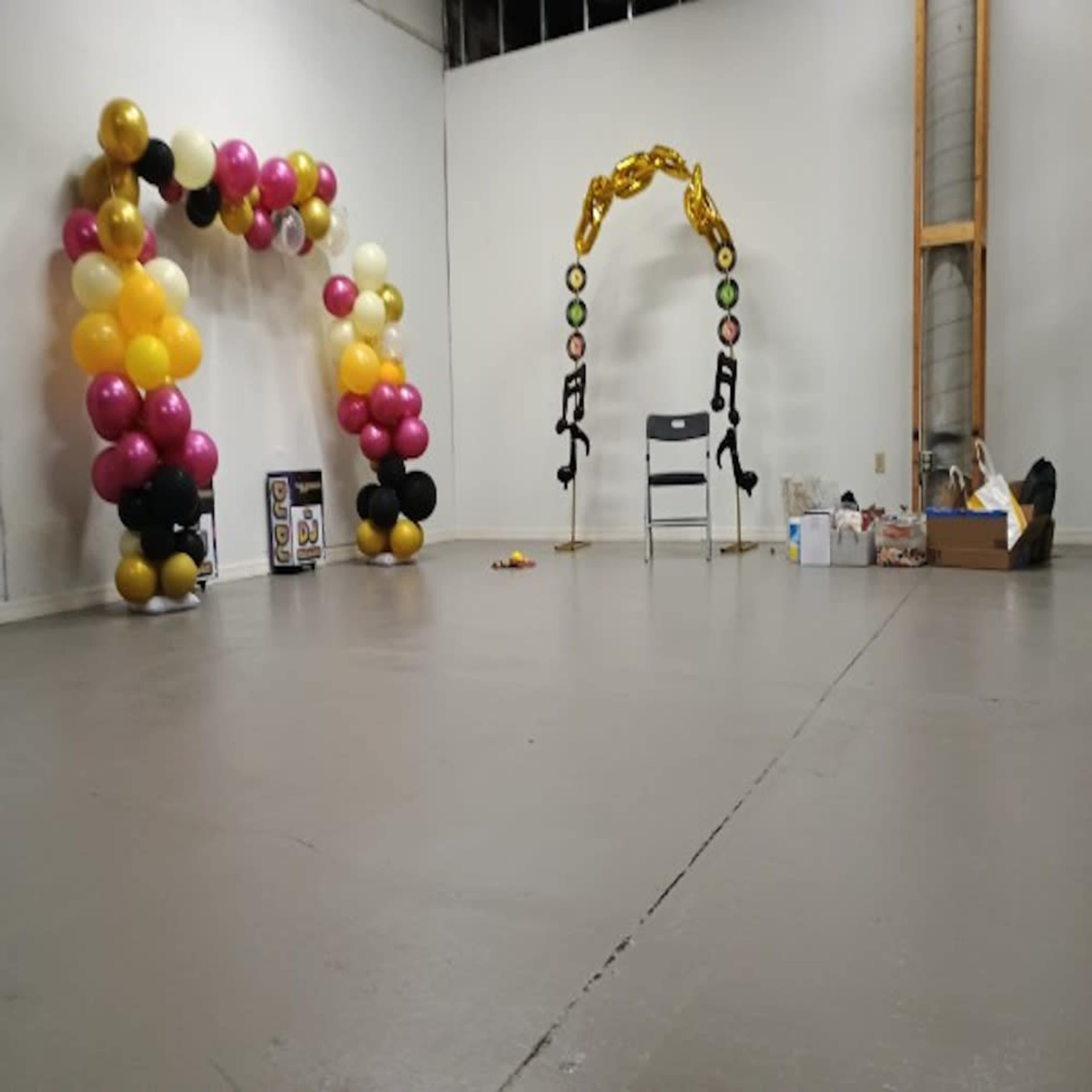 The scene features a large, empty room with two balloon arches—one with pink and black balloons, and the other with yellow and black balloons—alongside a single chair and some scattered items.