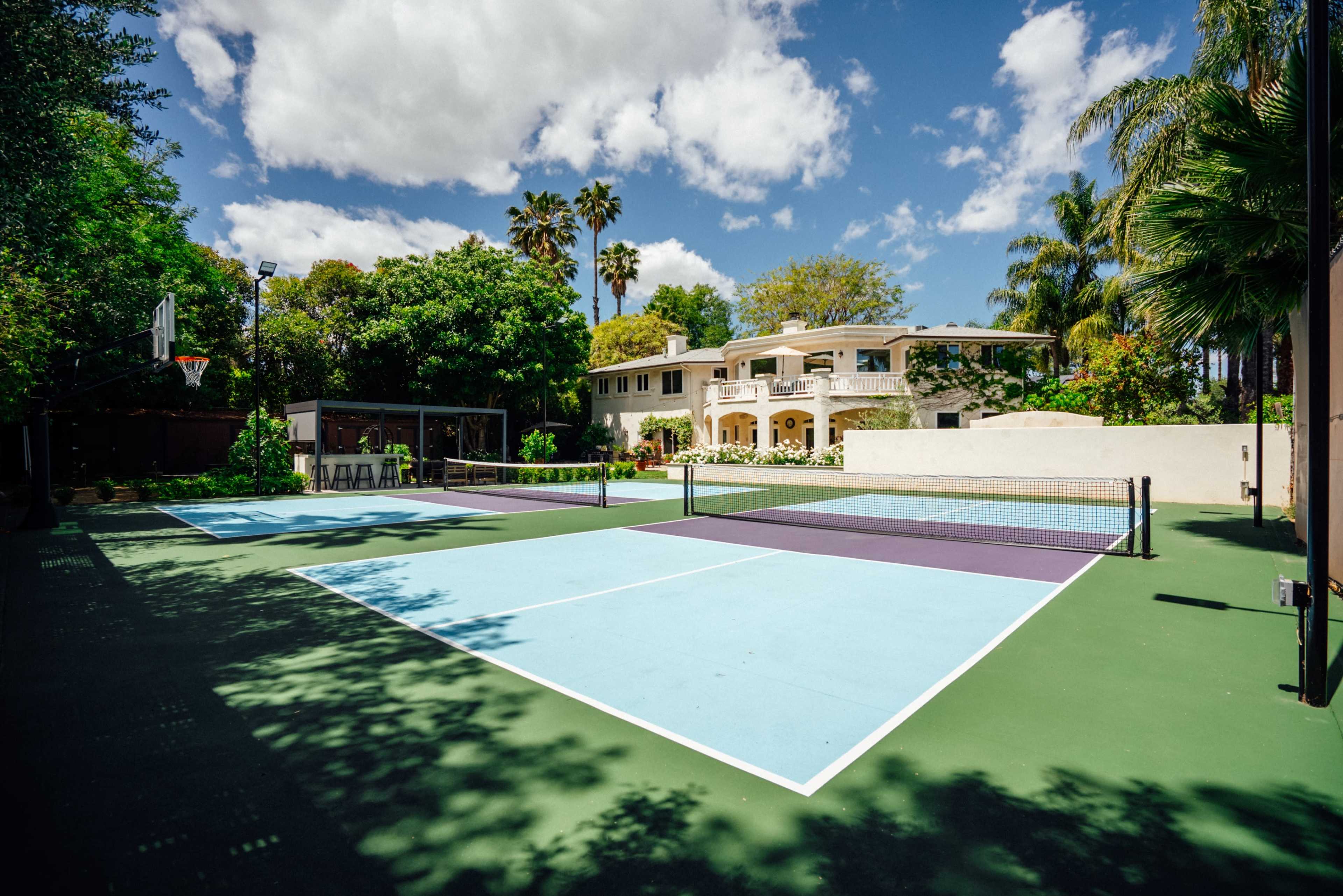 The image shows a private outdoor sports court, featuring two tennis courts and a basketball hoop, surrounded by lush greenery and a large house in the background.