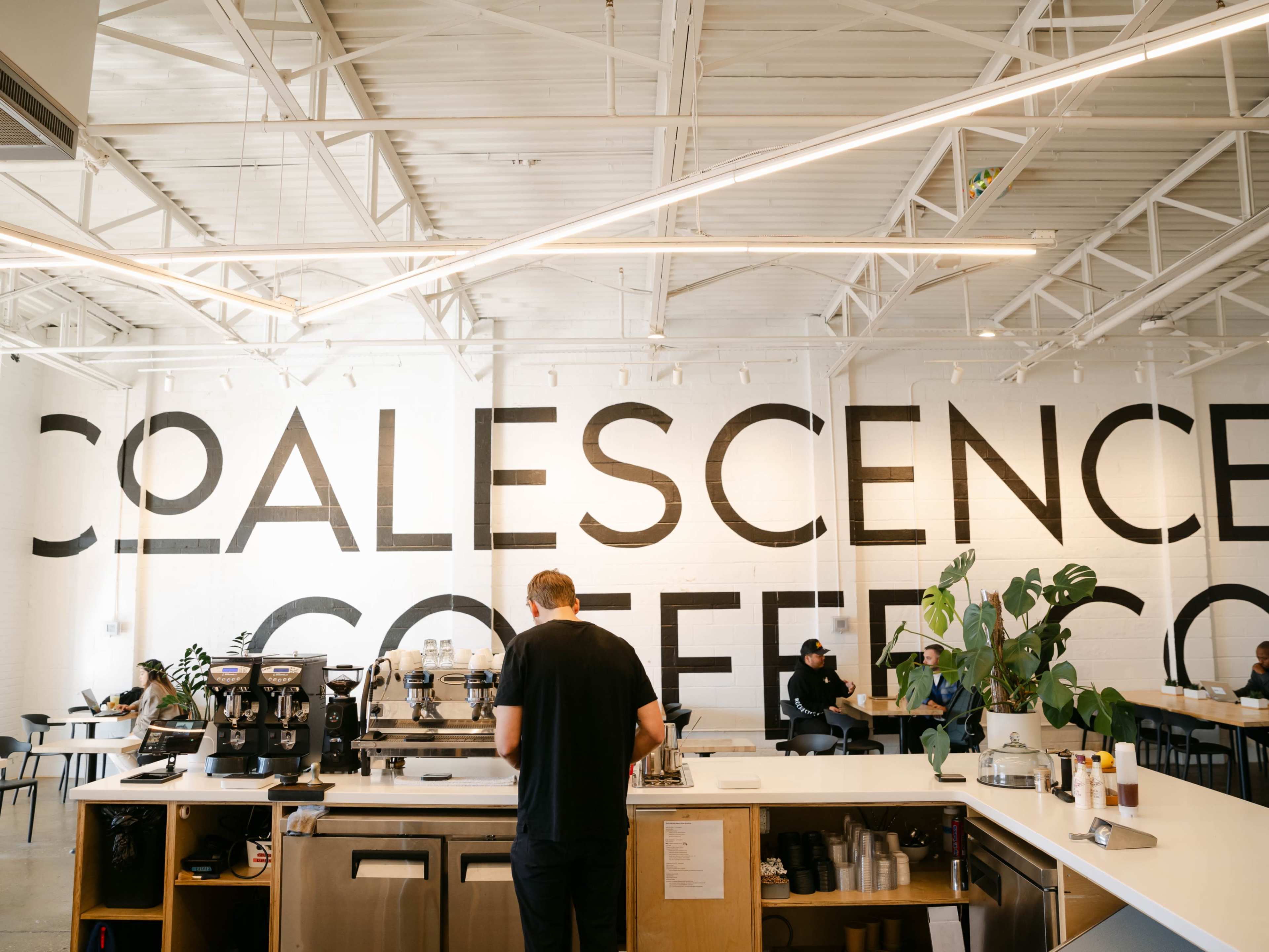A coffee shop with a large black-and-white mural reading "COALESCENCE COFFEE" on the wall and patrons seated at tables.
