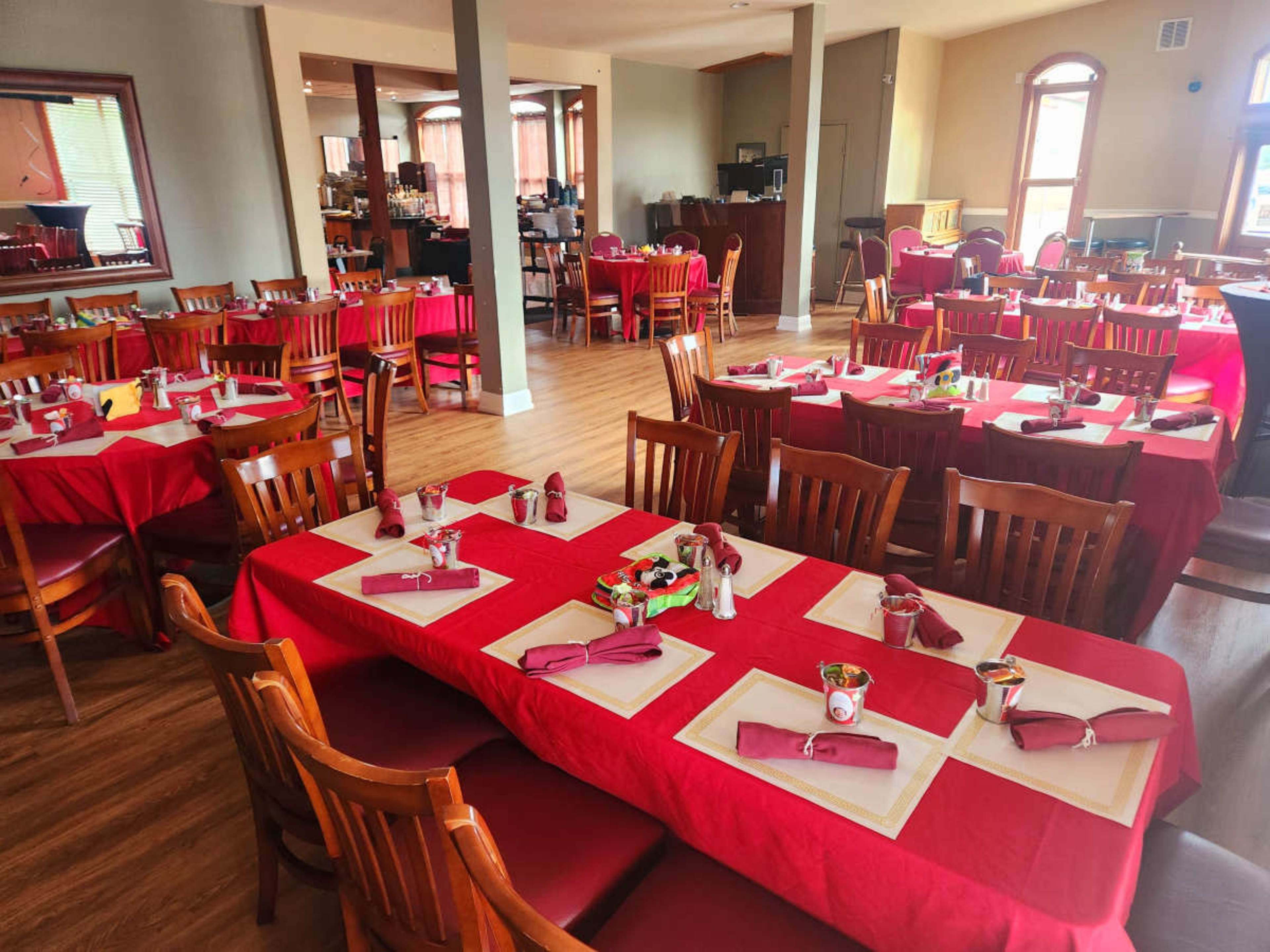 A restaurant dining area with multiple tables covered in red tablecloths, set with utensils and condiments.