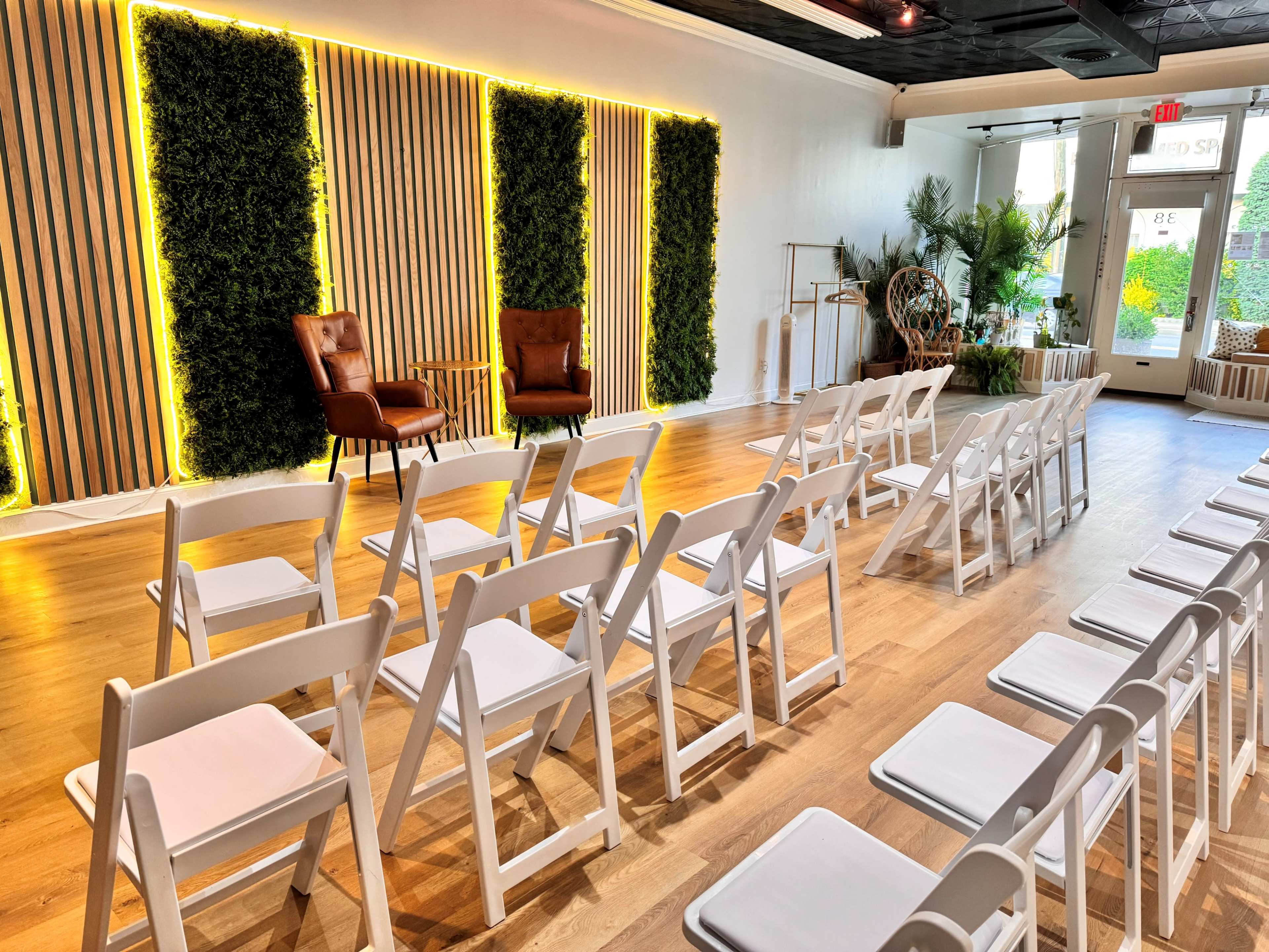 A neatly arranged seating area with white chairs facing two brown armchairs against a backdrop of green wall panels illuminated by soft lighting.