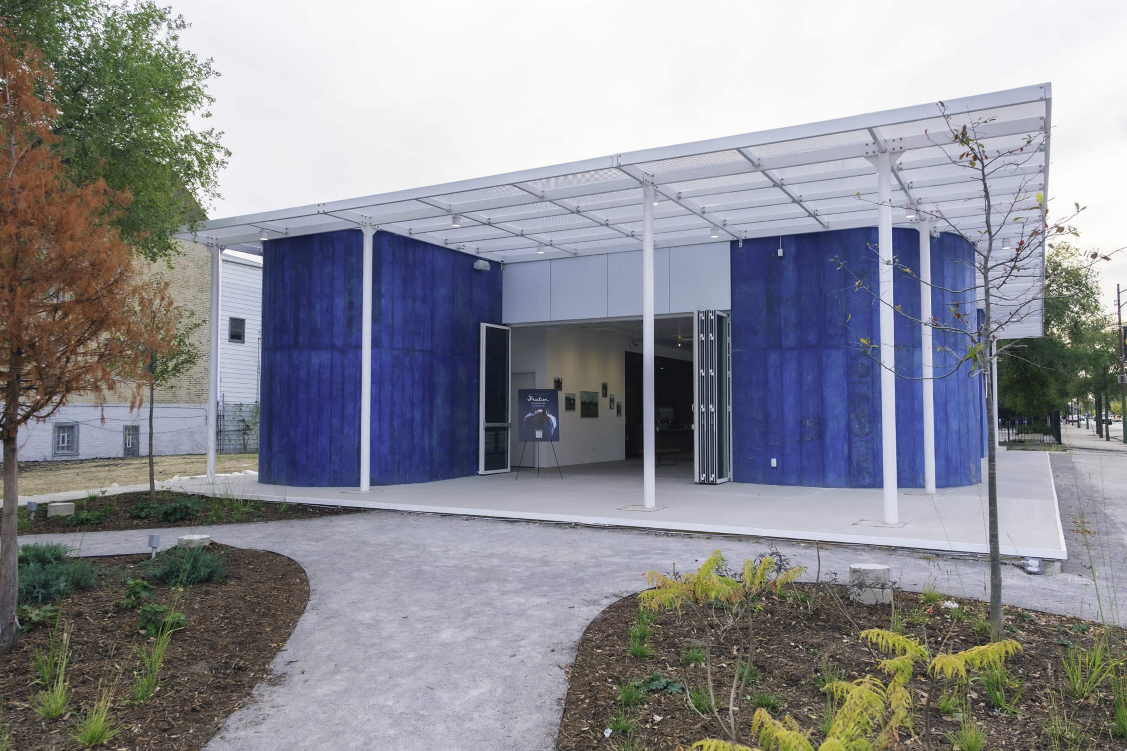 The image shows a modern blue building with a glass roof, situated on a landscaped path with plants and trees.
