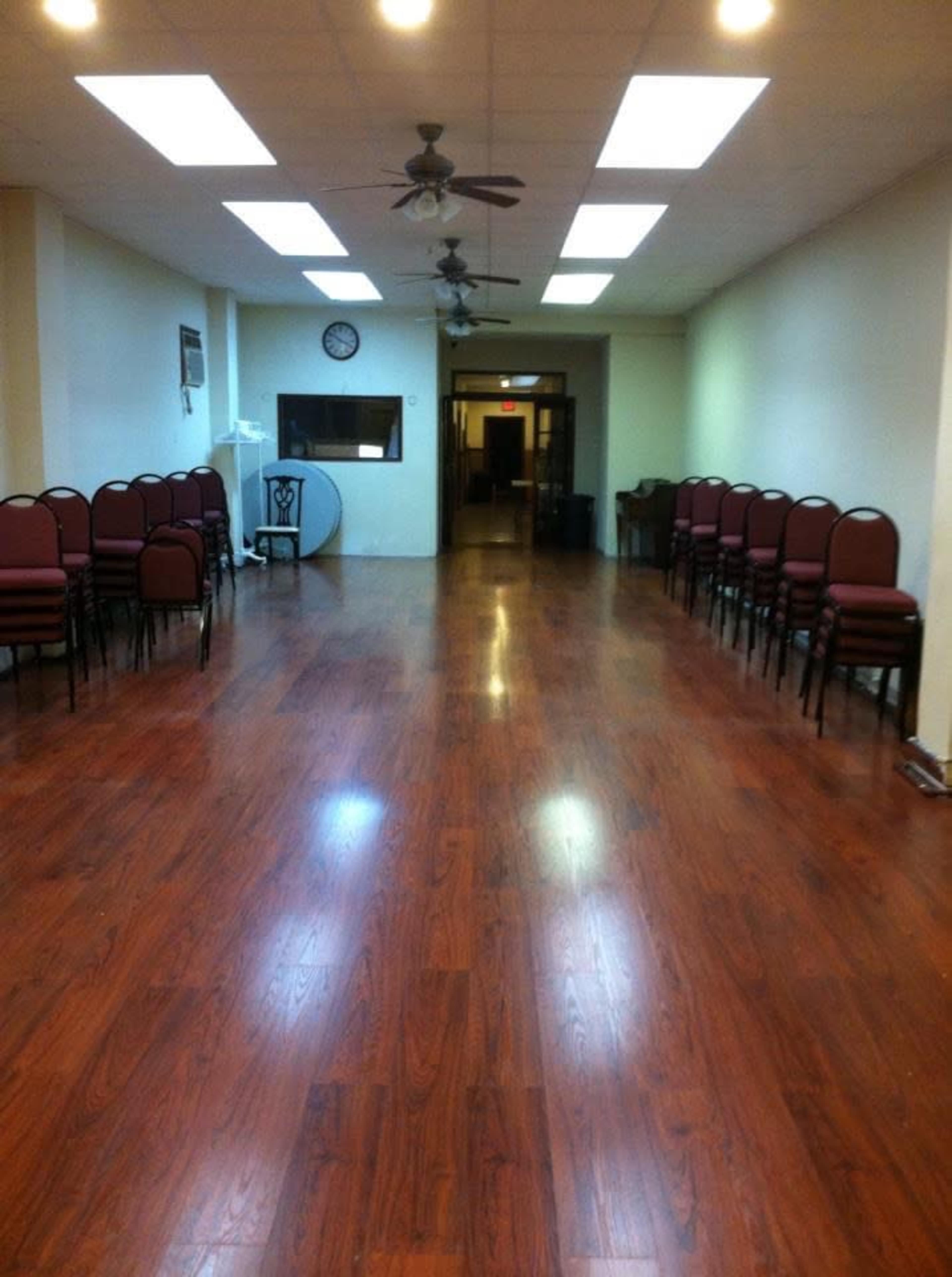 A spacious, empty room with wooden flooring, lined with red chairs on one side, and a clock and an adjacent entrance visible at the far end.