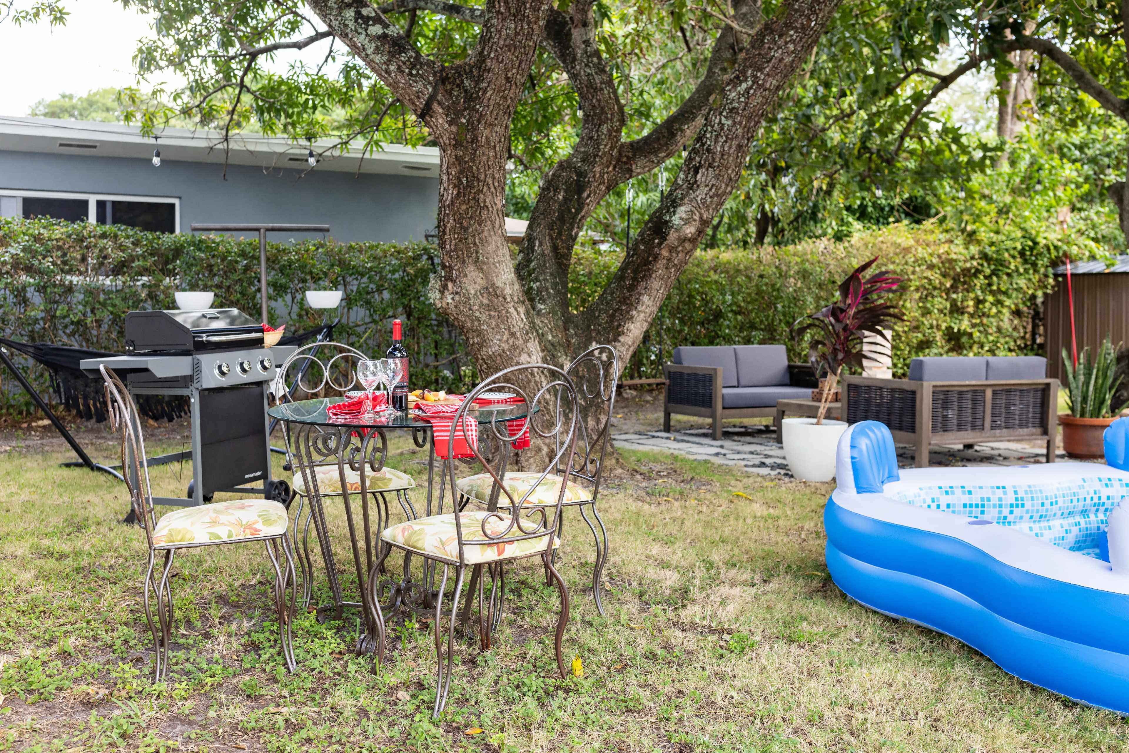 A backyard setup features a grill, a table with chairs, a small inflatable pool, and lounge seating beneath a tree.