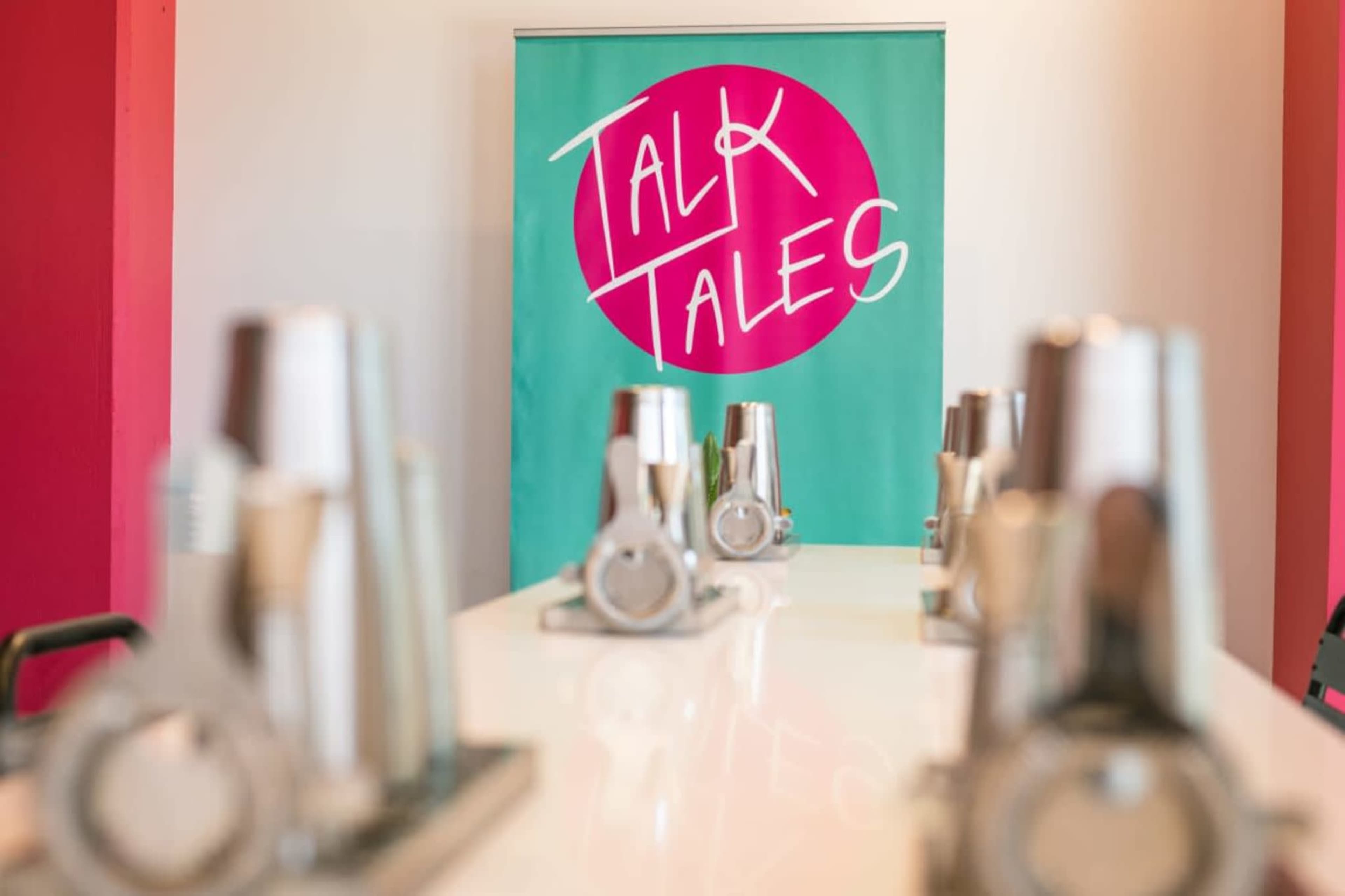 The image shows a bright, modern room with a banner reading "TALK TALES" and several metallic objects lined up on a table.