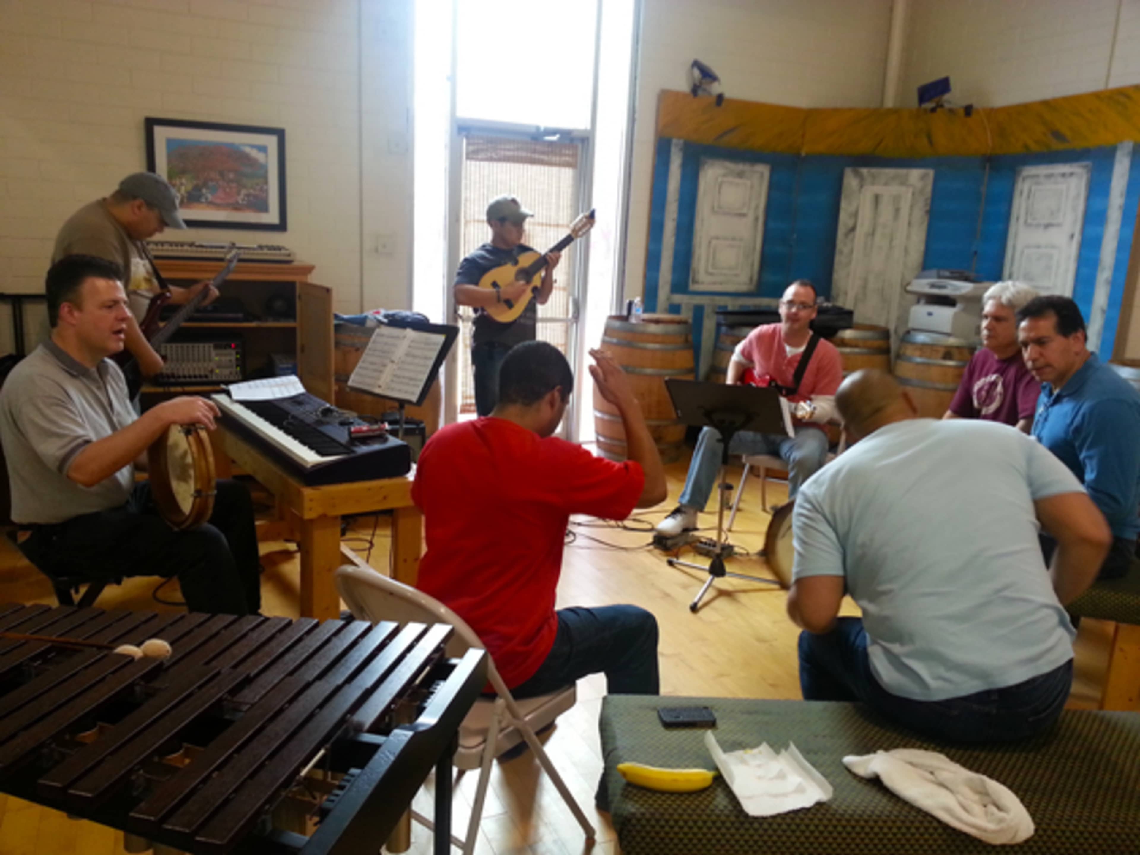 A group of musicians is gathered in a room, playing various instruments, while some are seated and engaged in conversation.