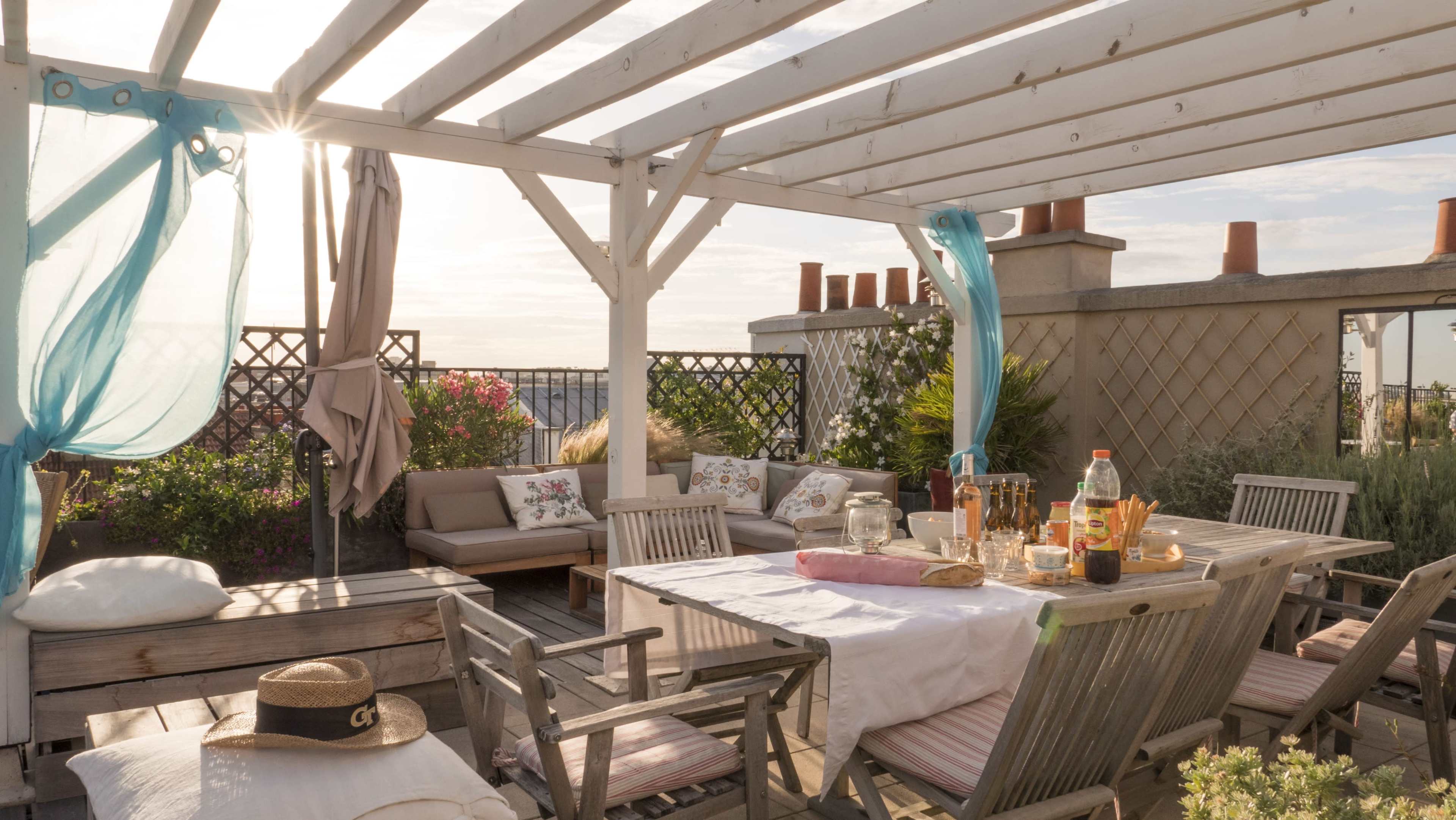 A spacious rooftop terrace features a wooden dining table, comfortable seating, and decorative plants under a pergola with flowing curtains.