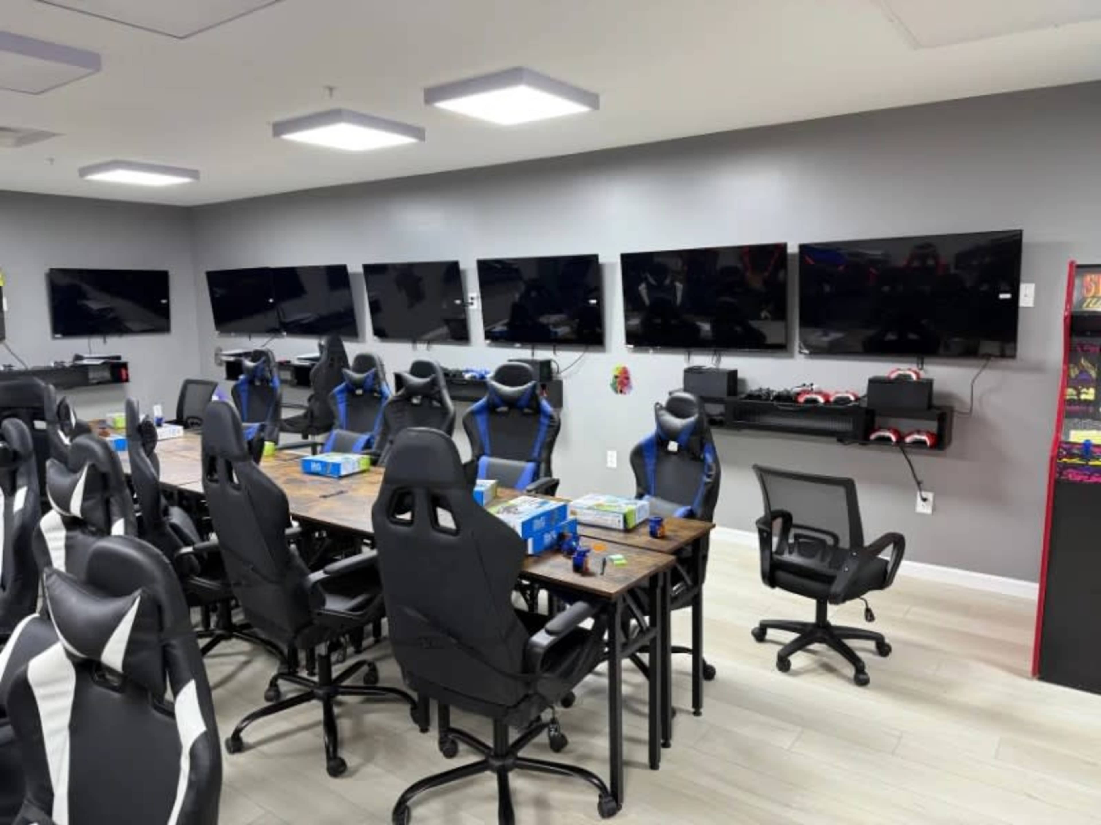 The image shows a modern conference room with multiple television screens mounted on the walls, a long table surrounded by black office chairs, and gaming equipment on shelves.