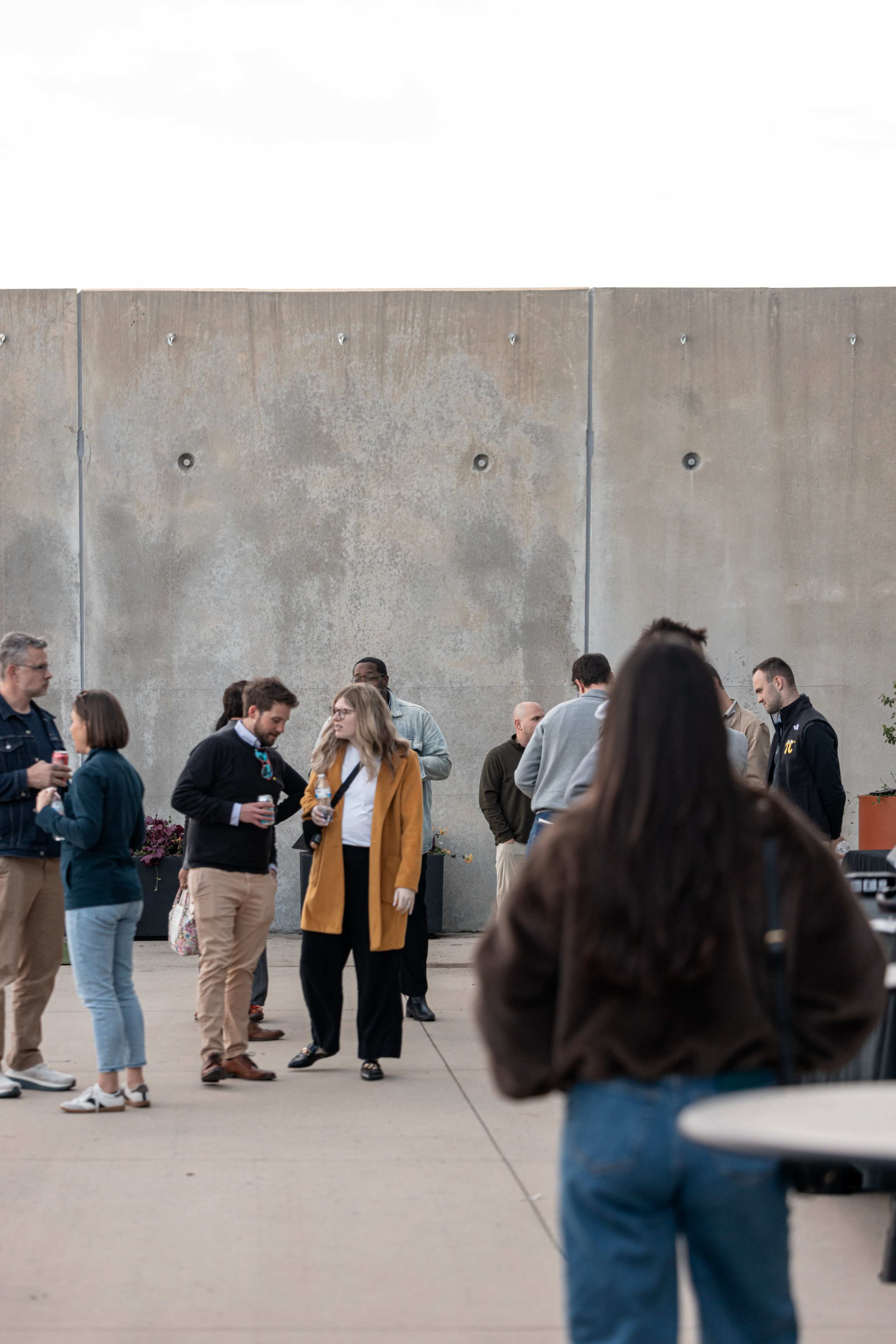A group of people are gathered outdoors on a concrete patio, some holding drinks and engaged in conversation.