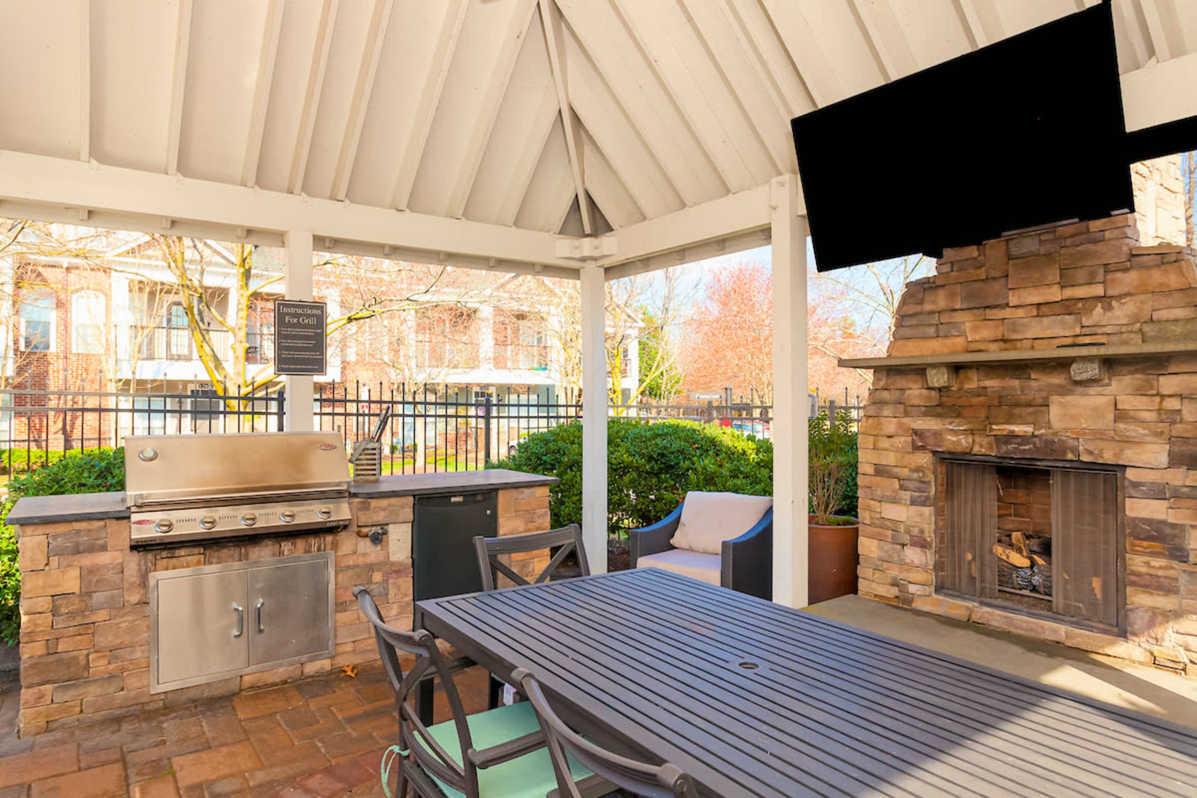 An outdoor grilling area with a stone fireplace, a built-in metal grill, and a wooden dining table beneath a covered pavilion.