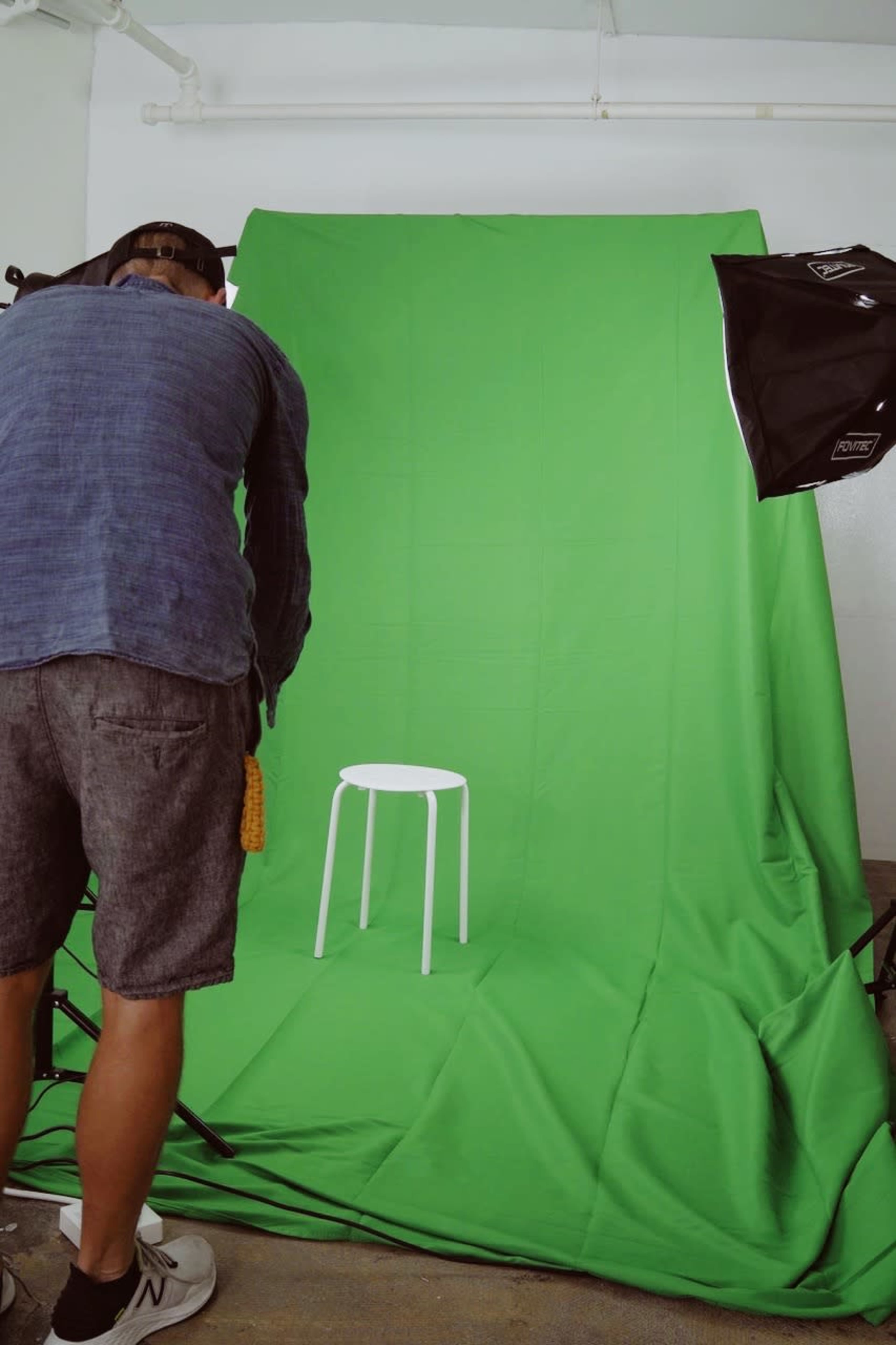 A person adjusts equipment in front of a green backdrop with a white stool positioned in the center.