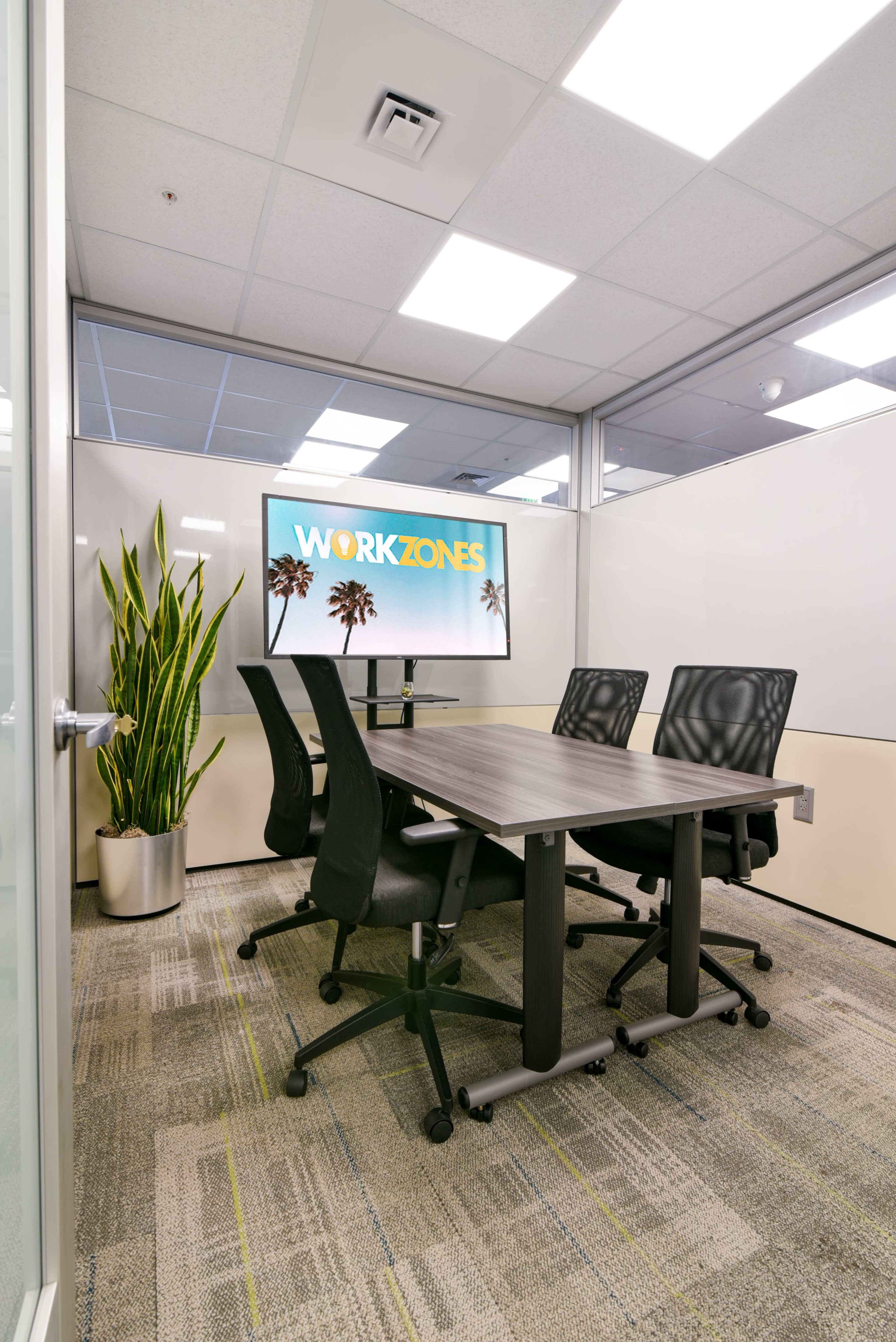 A conference room features a wooden table, four black swivel chairs, a large screen displaying "WORKZONES," and a tall plant in a pot.