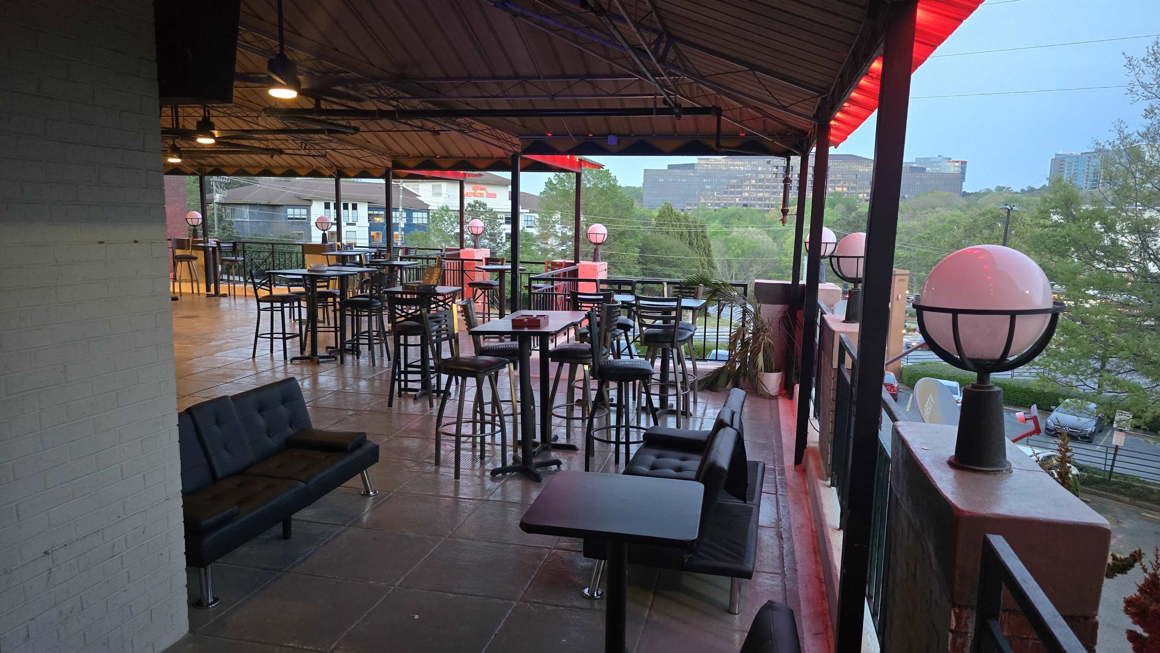 The image shows an outdoor terrace area of a bar or restaurant with black seating, small tables, and decorative lighting.