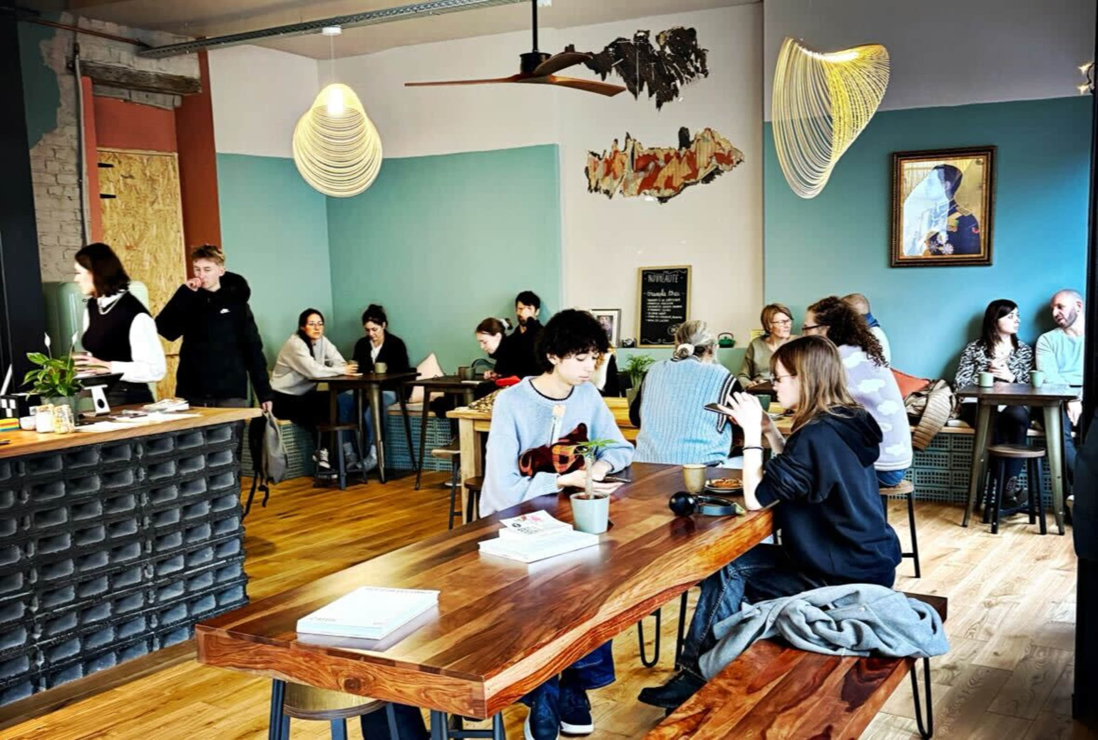 The image shows a cafe with patrons seated at various tables, some engaging in conversation while others work on laptops, surrounded by modern decor and plants.