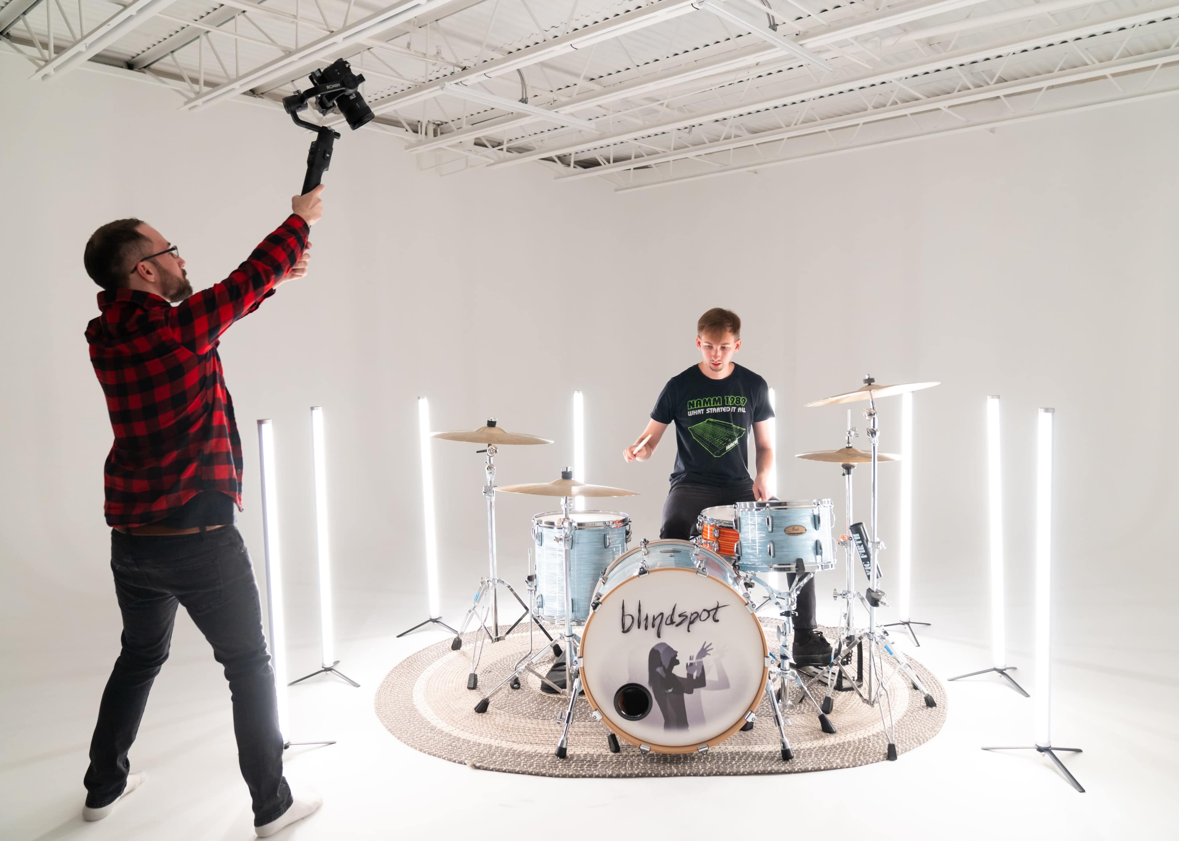 A drummer plays on stage while a man holds a camera on a stabilizer above him in a well-lit studio.