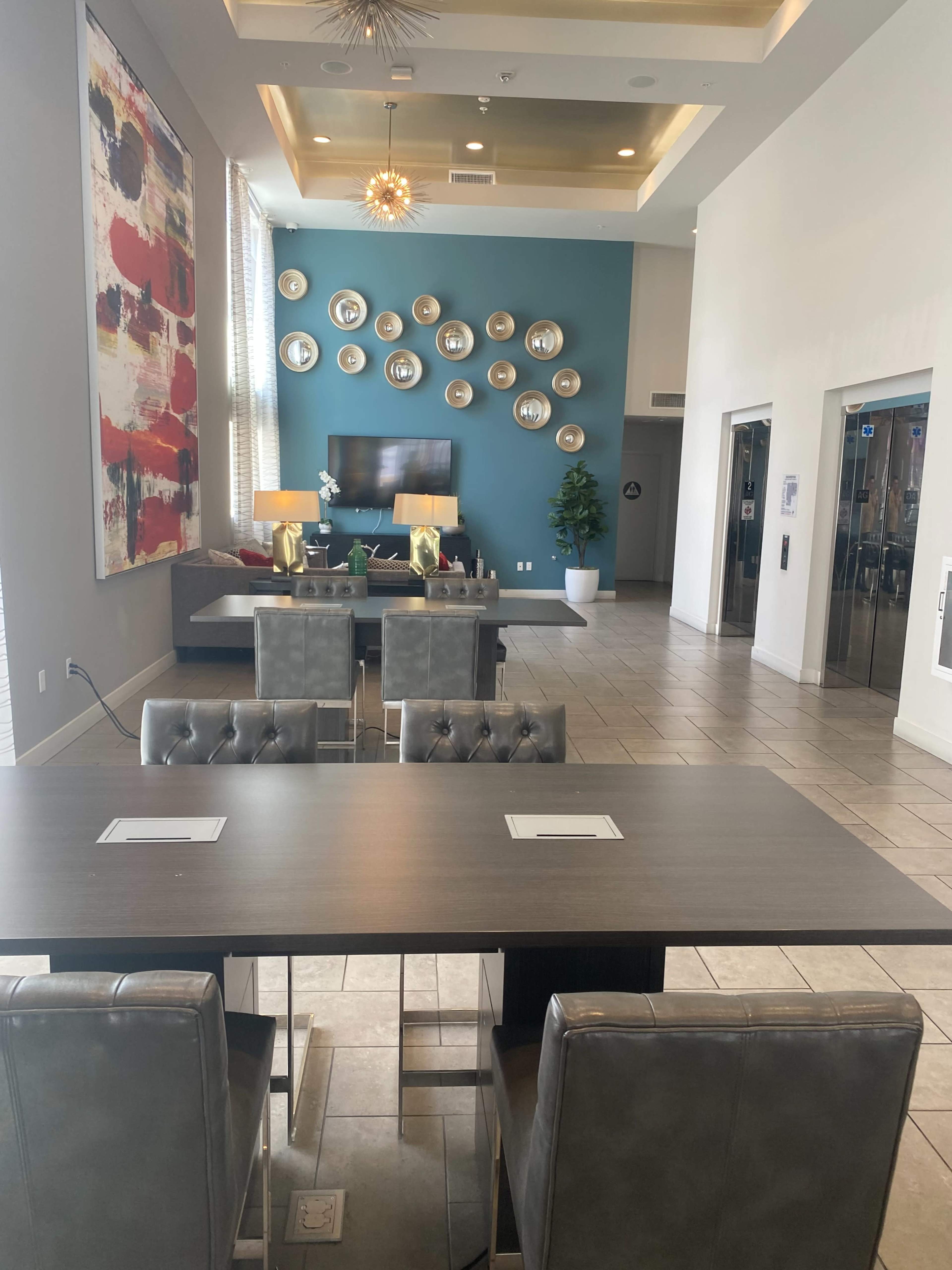 A modern lobby area with a large table and chairs, a seating arrangement, elevators, and decorative wall art.