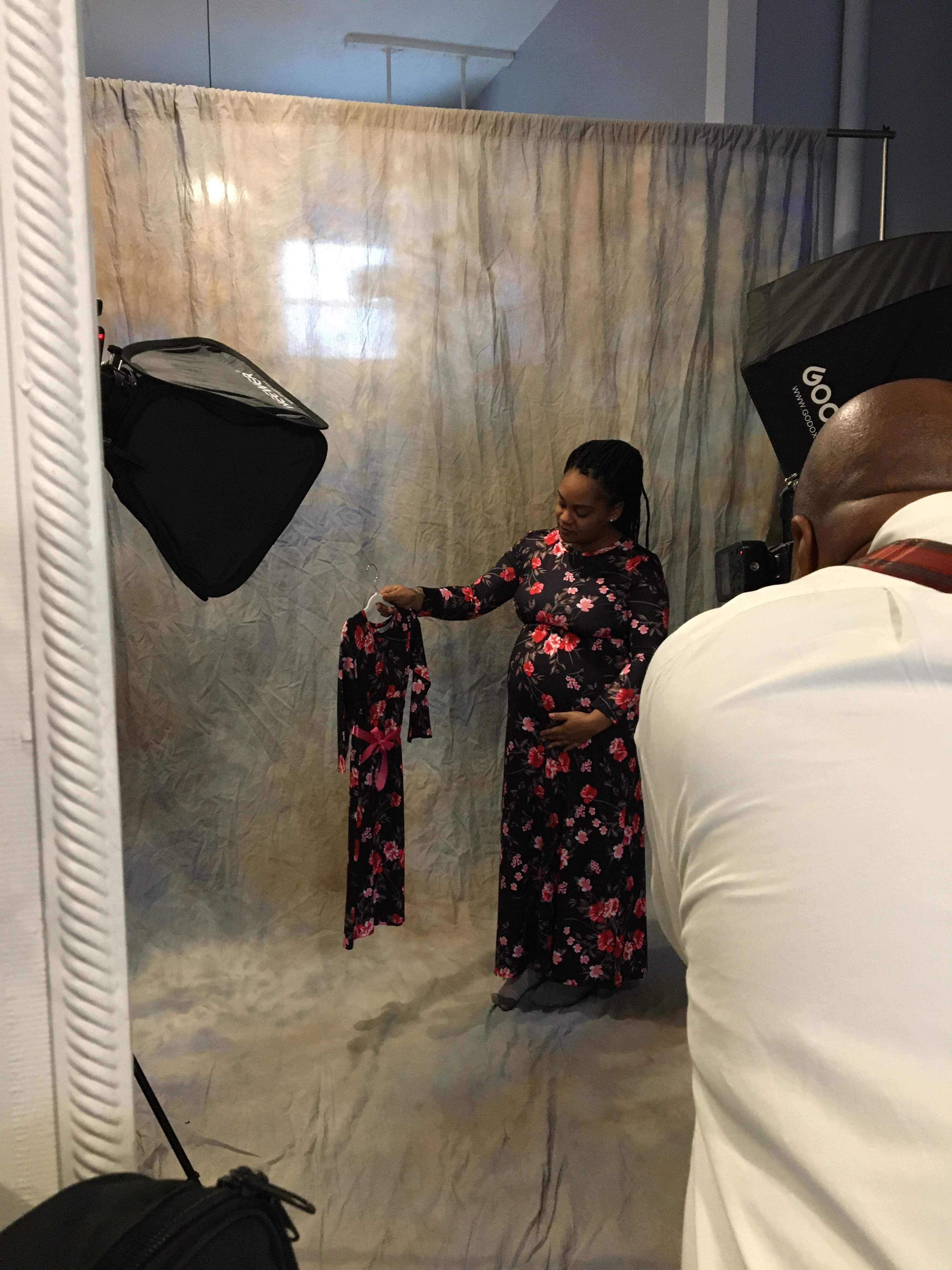 A pregnant woman holds a dress while posing for a photo in front of a draped fabric background and studio lights.