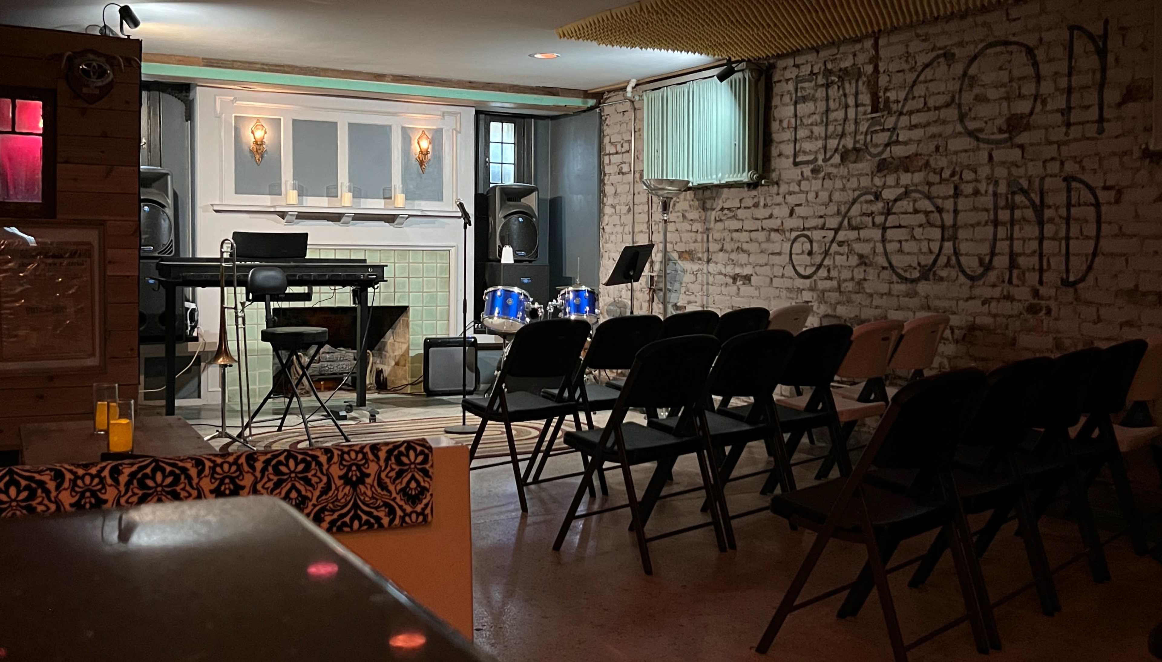 A small, empty performance space featuring a keyboard, drums, and rows of folding chairs arranged for an audience.