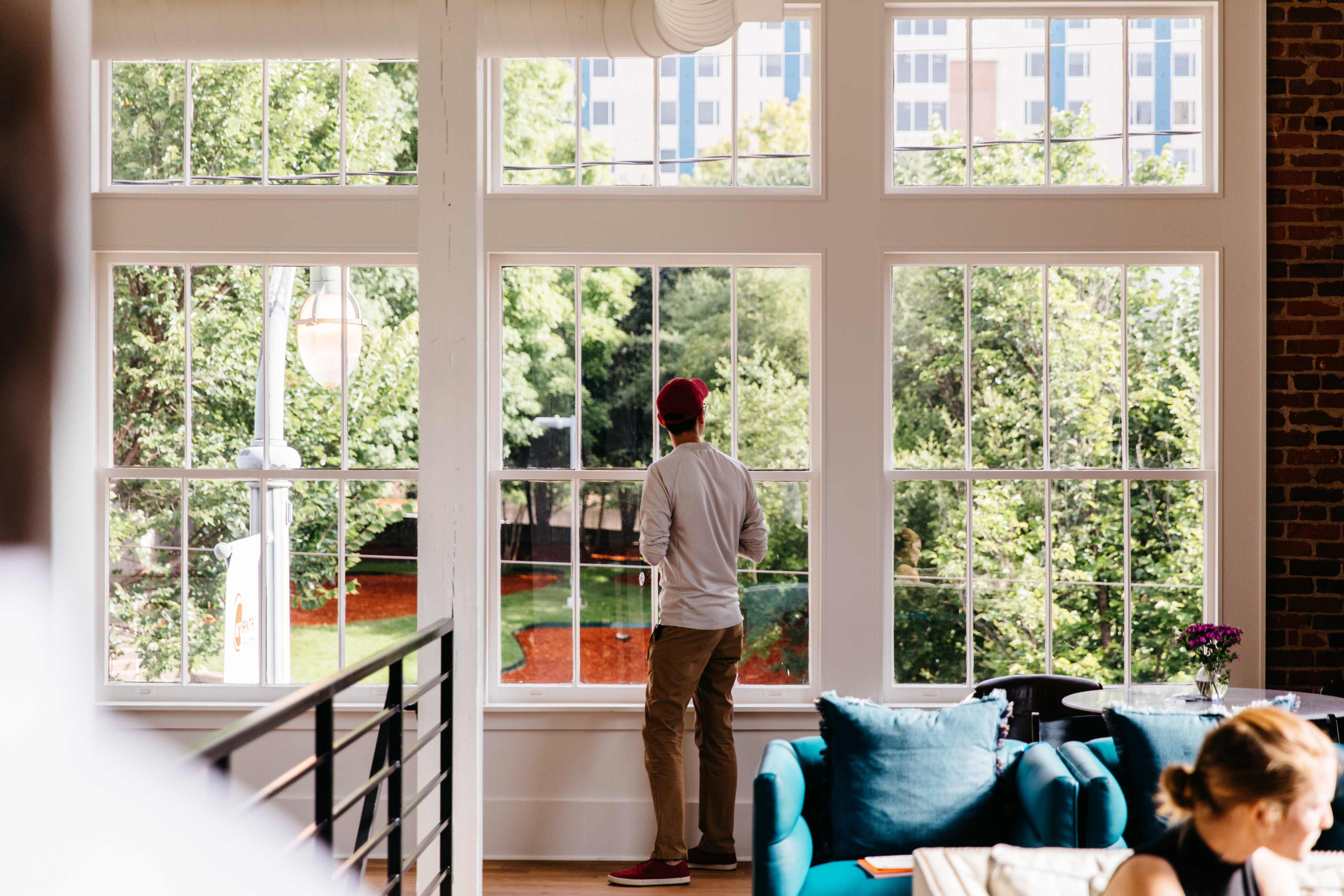 A person stands by a large window, looking out at a green park area with buildings in the background.