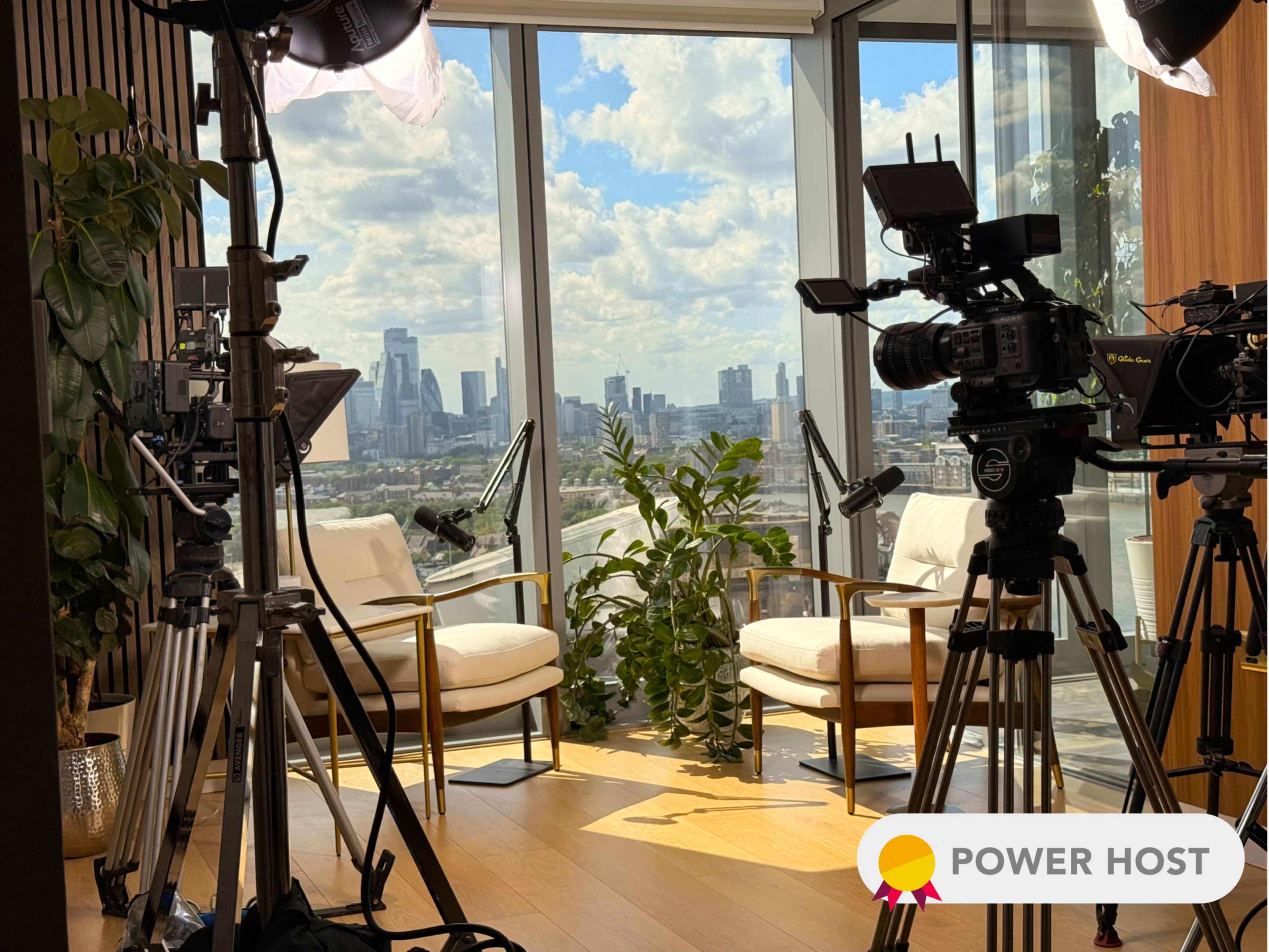The image shows a studio setup with two light-colored chairs facing a large window that offers a view of a city skyline, surrounded by video cameras and lighting equipment.