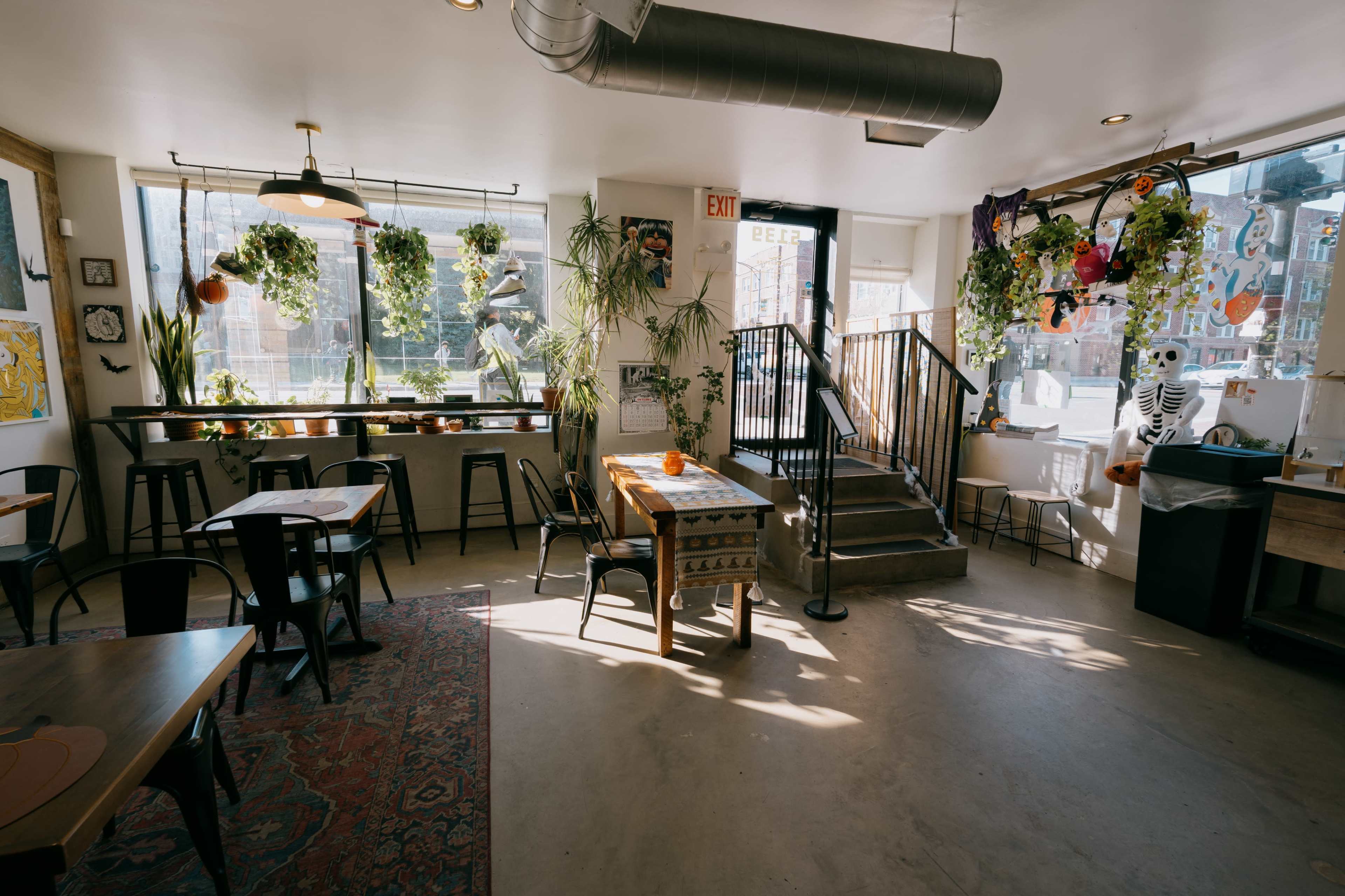 The image shows a cozy café interior with wooden tables, black metal chairs, and large windows adorned with plants.
