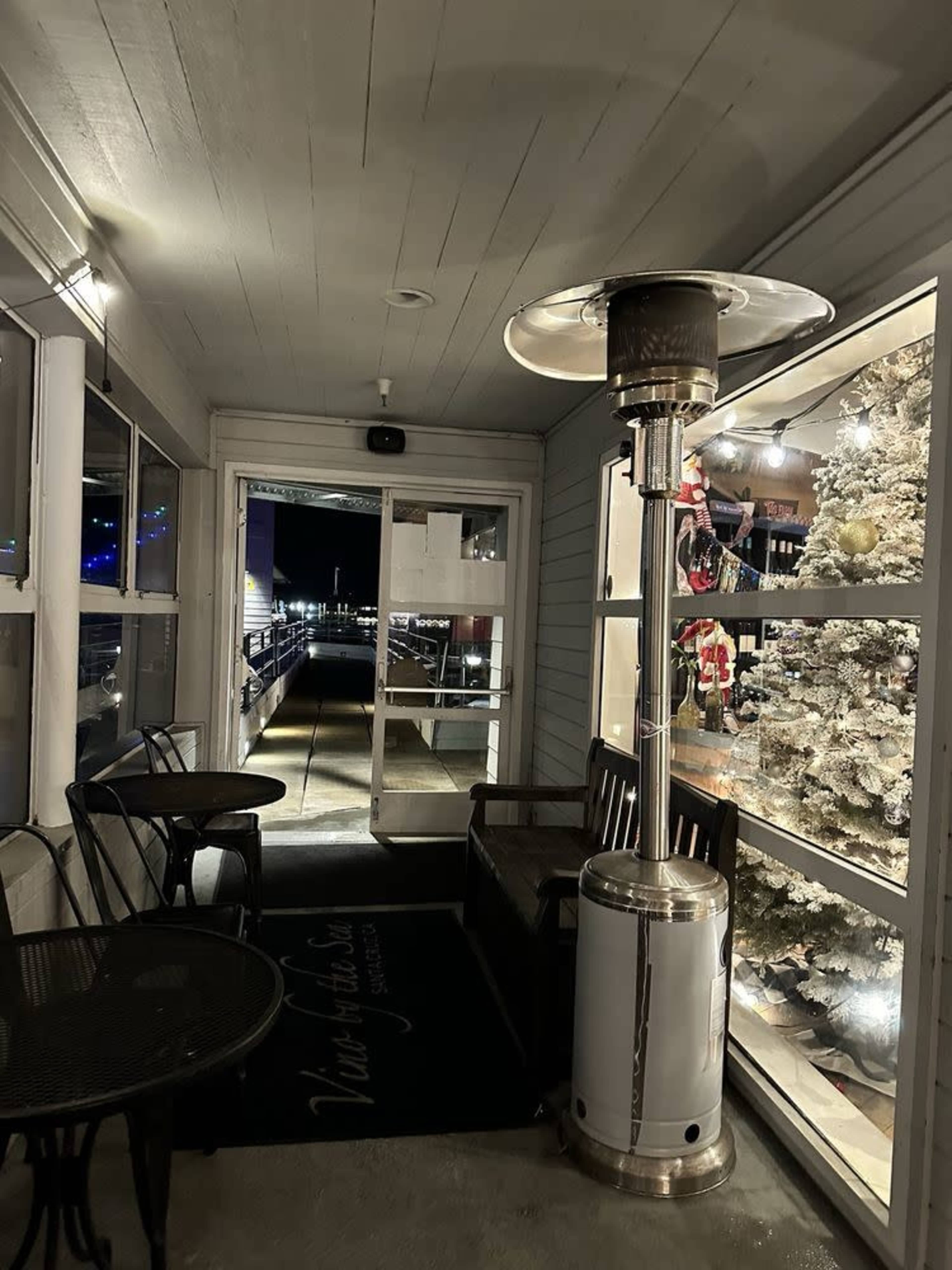 The image shows a covered porch area with a patio heater, two tables, and a bench, leading to a glass-paneled storefront decorated for the holidays.