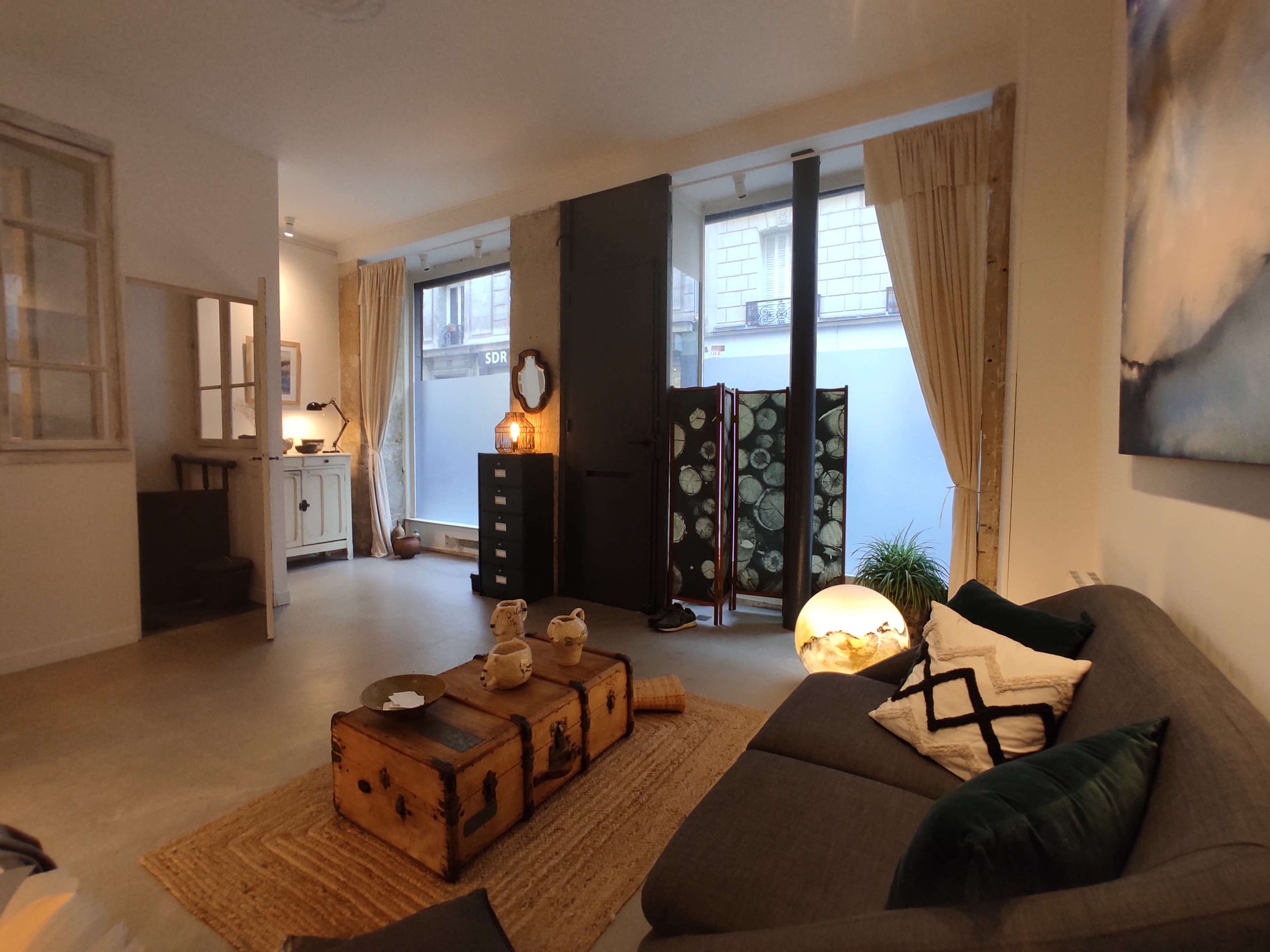 The image shows a modern living room featuring a gray sofa, a pair of wooden crates as a coffee table, and large windows with sheer curtains, illuminated by natural light.