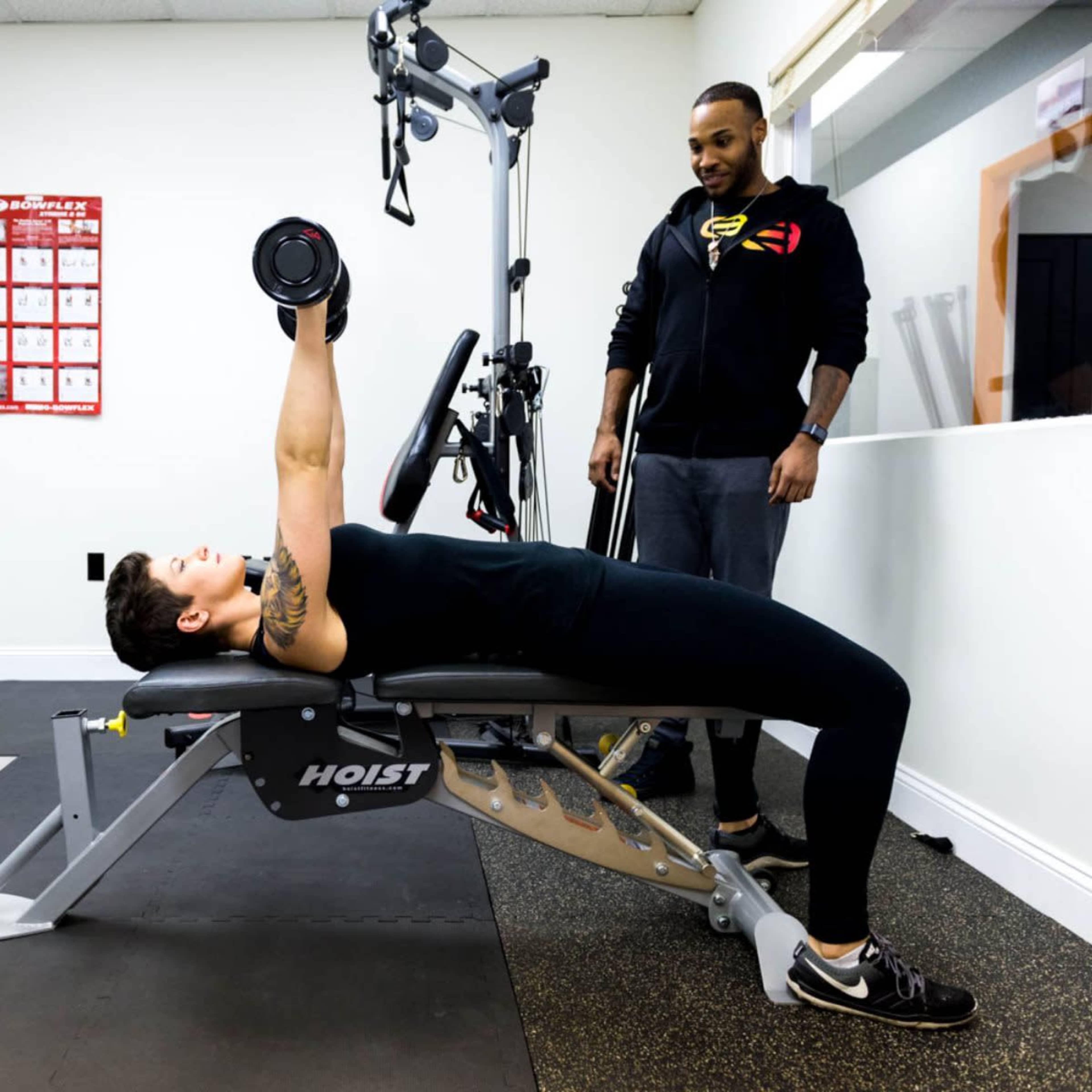 A person lies on a bench performing a dumbbell chest press while a trainer observes nearby in a gym setting.