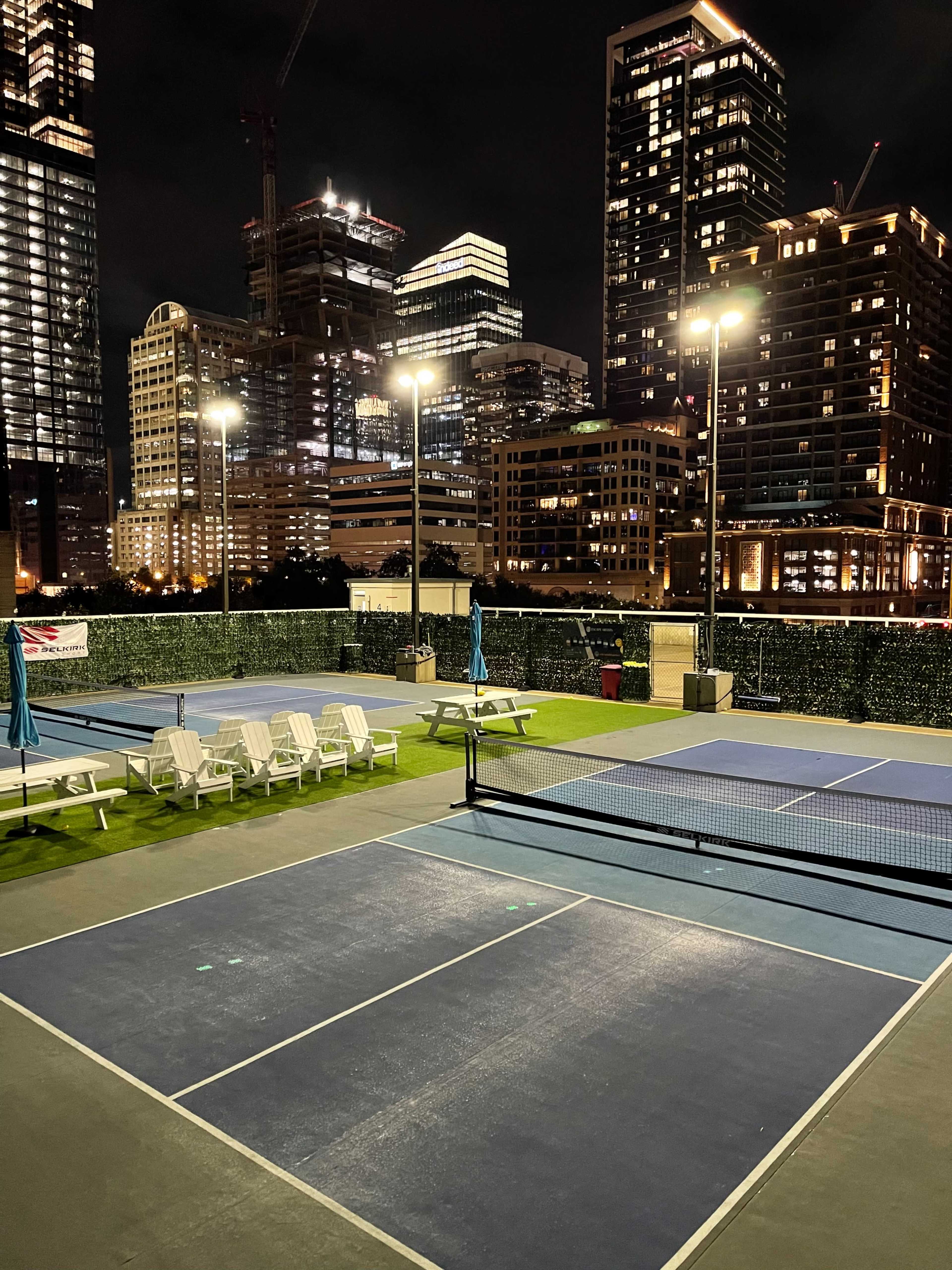 An empty tennis court at night, surrounded by lit buildings in an urban setting.