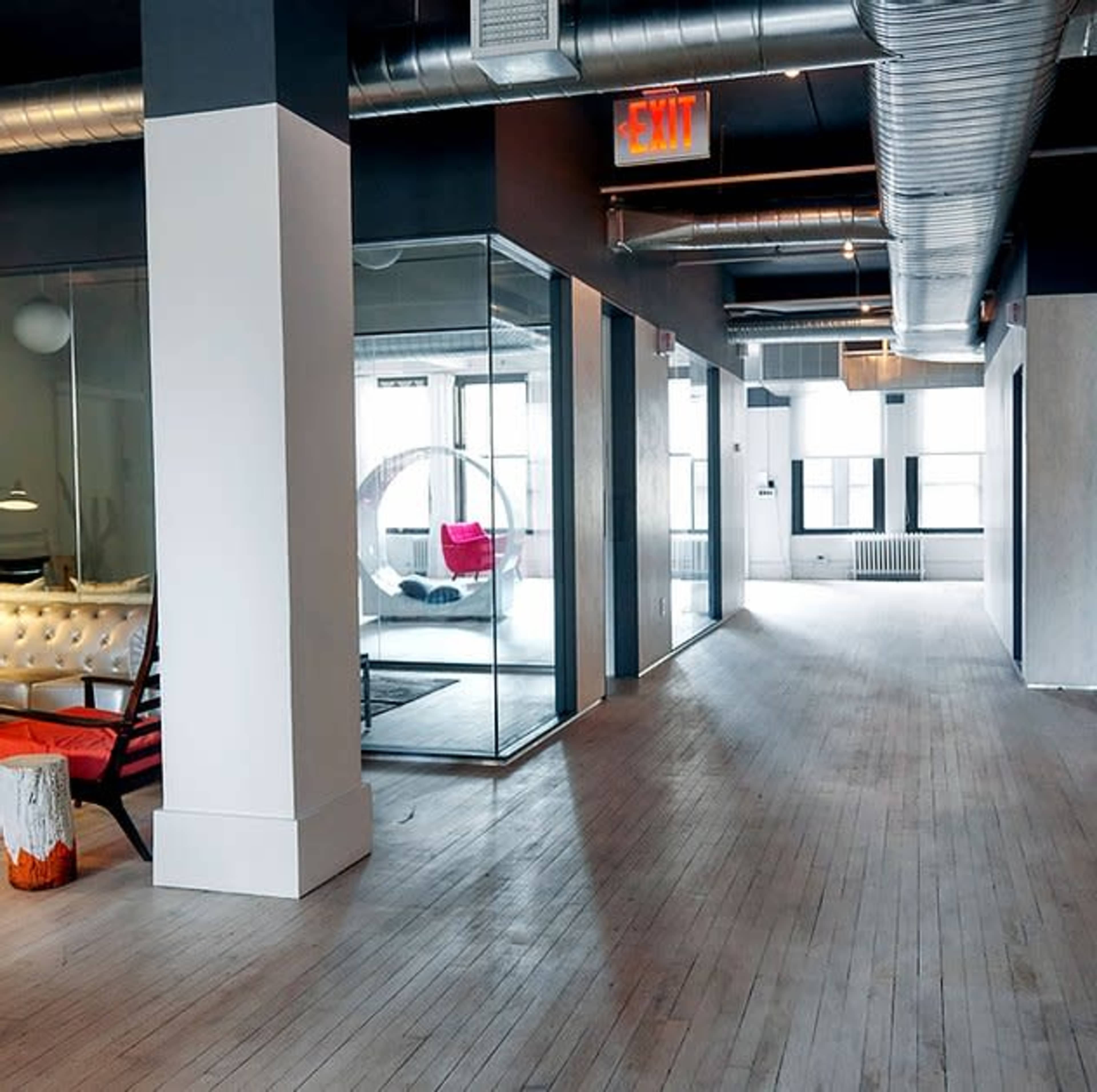 The image shows a spacious, modern office interior with large windows, wooden flooring, glass-walled meeting rooms, and a clear exit sign.